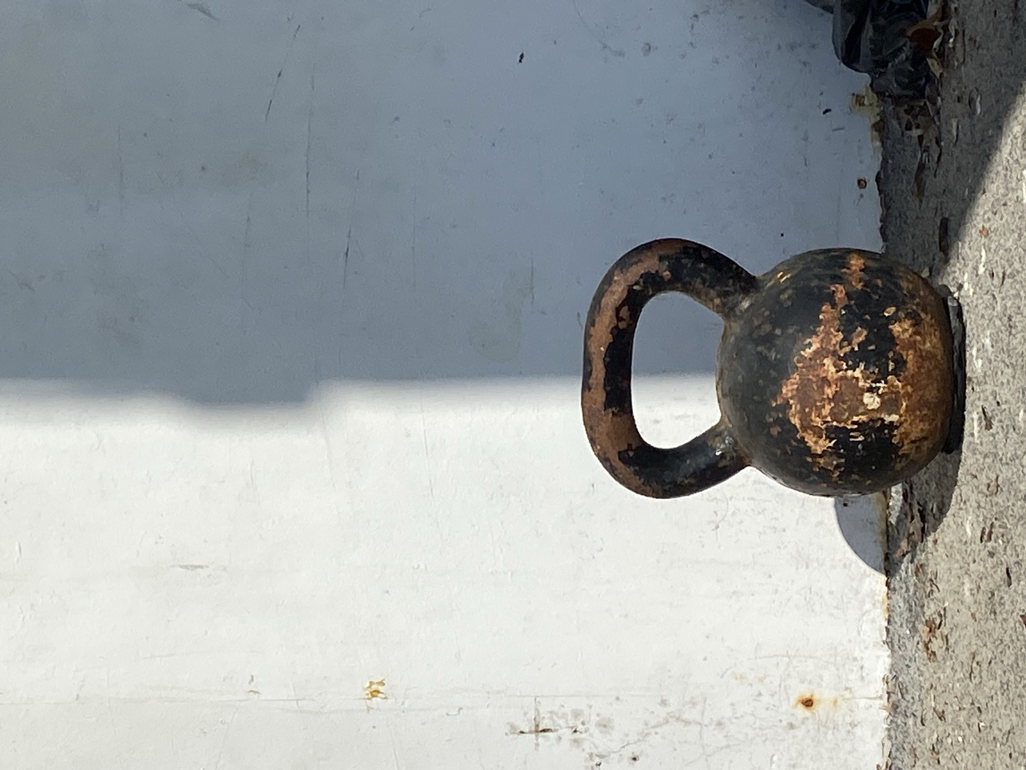The image depicts an old, rusted kettlebell sitting on a concrete surface, potentially a sidewalk or ledge. The kettlebell is black with patches of exposed rust. A section of a white painted wall is visible behind the kettlebell. A small portion of black trash bag is visible on top of the kettlebell and to the upper right. The photograph is taken in bright daylight, casting a shadow from the kettlebell onto the wall. The location is Prijevor, Montenegro.