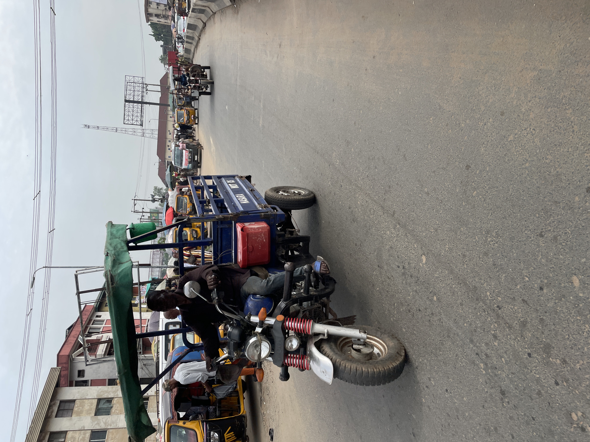 The image shows a street scene in a city. In the foreground, a man is riding a three-wheeled motorized vehicle, which appears to be a type of auto rickshaw or tuk-tuk, with a green canopy and a blue cargo box. The vehicle is moving on a paved road. In the background, other vehicles, including more auto rickshaws and cars, are visible. Buildings line the street, and power lines are overhead. The sky is overcast. The scene appears to be a busy urban environment, possibly a market area or a transportation hub.