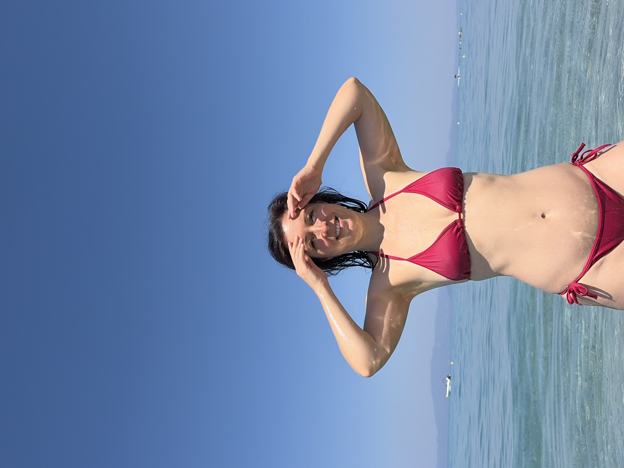 A woman in a dark red bikini stands in the clear, shallow water of a body of water, likely the sea, given the location context of Pallini Municipal Unit, Greece. The sky is a vibrant, clear blue, indicating it is daytime, likely sunny. The woman is looking towards the camera with a slight smile, her hands raised to her forehead, perhaps shielding her eyes from the sun. Her hair is dark and appears wet. The water is calm and translucent, with a hint of green tint, allowing the seabed to be seen in some areas. In the distance, a few small boats or buoys are visible on the water. The overall mood is relaxed and summery, suggesting a vacation or leisure activity. There is no visible text in the image.