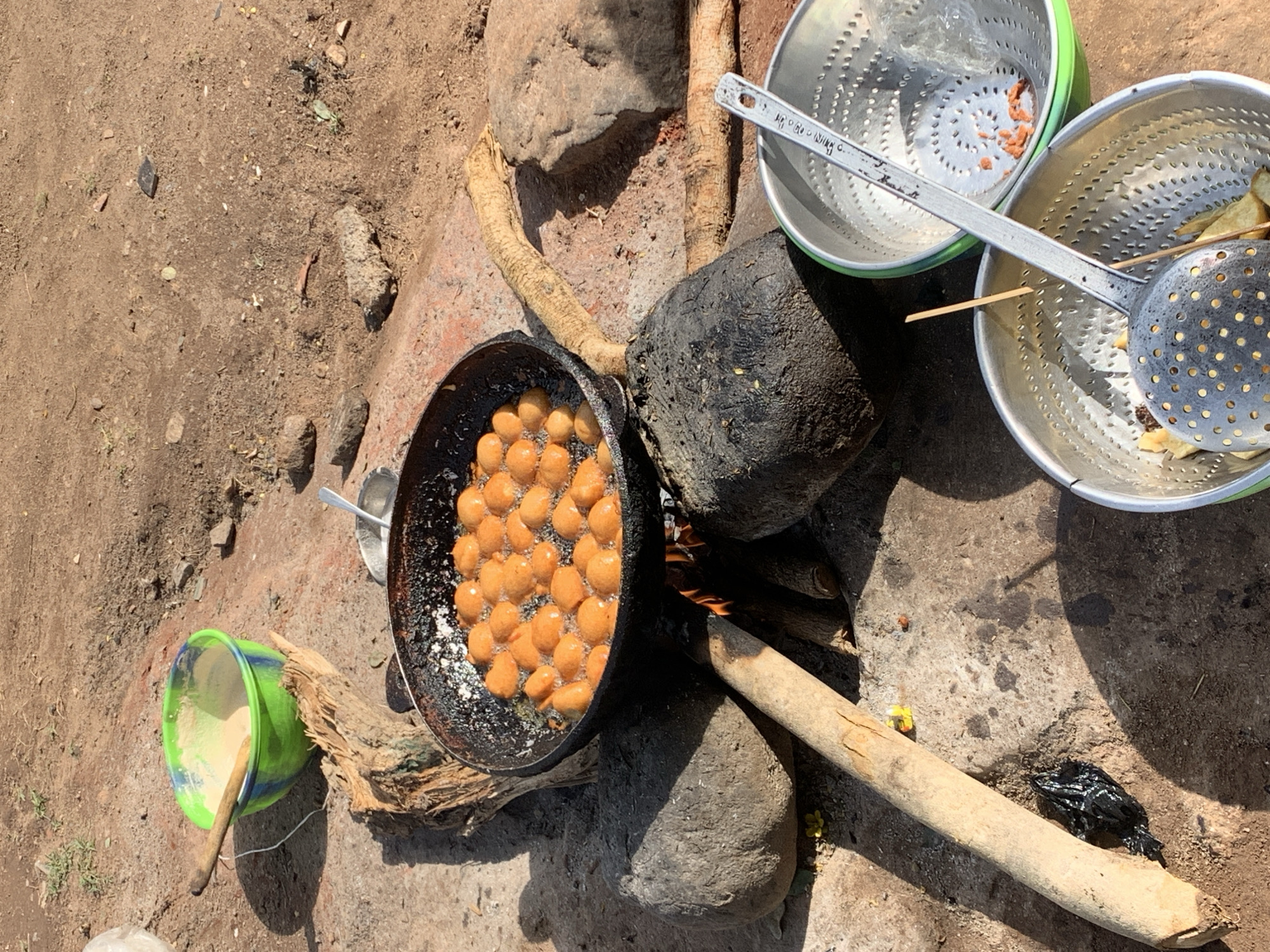 The image shows an outdoor cooking scene in Kabong, Nigeria. A black frying pan filled with spherical, golden-brown food items, likely fried dough, sits atop a makeshift stove constructed from stones and pieces of wood. A fire is burning underneath the pan. Two silver colanders with green rims, one containing a metal skimmer, are positioned to the right of the stove. A green plastic bowl, containing a light-colored substance and a wooden spoon, rests on the ground to the left. The ground is dirt with scattered rocks and debris. The scene appears to be in a rural or undeveloped area.