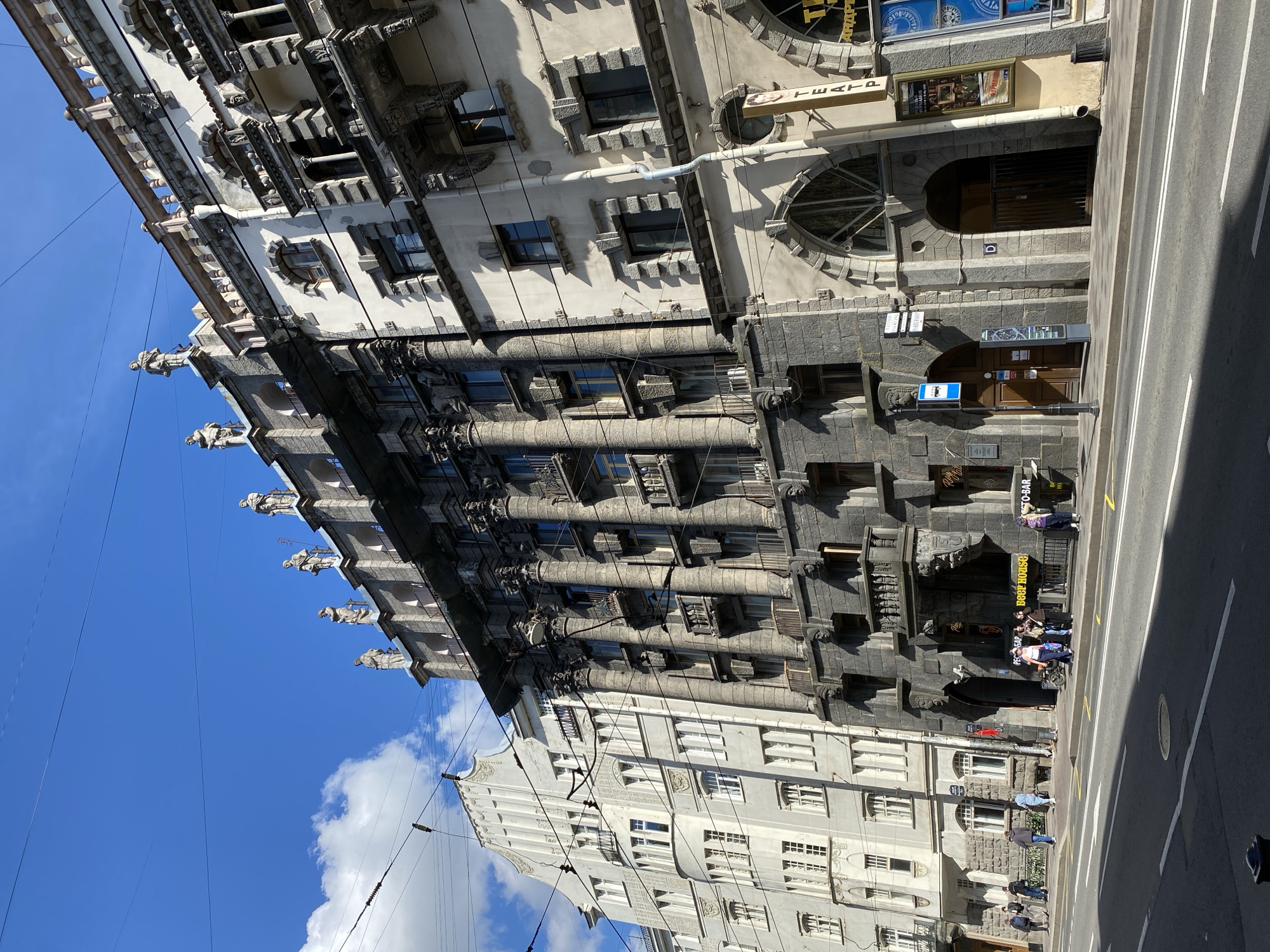 The image captures a vibrant street scene in Saint Petersburg, Russia, under a bright, clear blue sky with scattered white clouds, suggesting good weather during daytime.
The prominent subject is a multi-story building dominating the right side of the frame. It features a highly distinctive and ornate architectural style, likely Art Nouveau. The lower levels are clad in rough-hewn, dark grey stone with heavily textured surfaces and deeply recessed windows. Intricate sculptural details and relief work adorn these lower floors. The upper stories transition to a lighter, possibly white or light grey, facade, maintaining elaborate window frames, decorative balconies, and arched elements. A notable feature is a series of large, gargoyle-like statues or figures protruding from the roofline. Overhead power lines crisscross the sky and buildings.
To the left of this grand structure, another multi-story building with a simpler, light grey facade and numerous windows can be seen, offering a visual contrast.
In the foreground, a paved asphalt road with clear white lane markings runs parallel to the buildings. A sidewalk separates the road from the building facades. Several people are visible on the sidewalk, walking individually or gathered in small groups, particularly near entrances to establishments. A bus stop sign is present near the ornate building.
Visible text on signs and posters includes:
* A vertical sign on the ornate building that reads "ТЕАТР" (Theatre).
* Below the theatre sign, a poster with the word "ПРЕМЬЕРА" (Premiere) is partly visible.
* A small number "53" is seen above an arched doorway.
* Signs on the lower level of the ornate building indicate "PESTO-BAR", "BEER KVASS", and one displays the year "1905".
The scene depicts a typical urban environment, showcasing historical architecture, street life, and the infrastructure of a modern city.