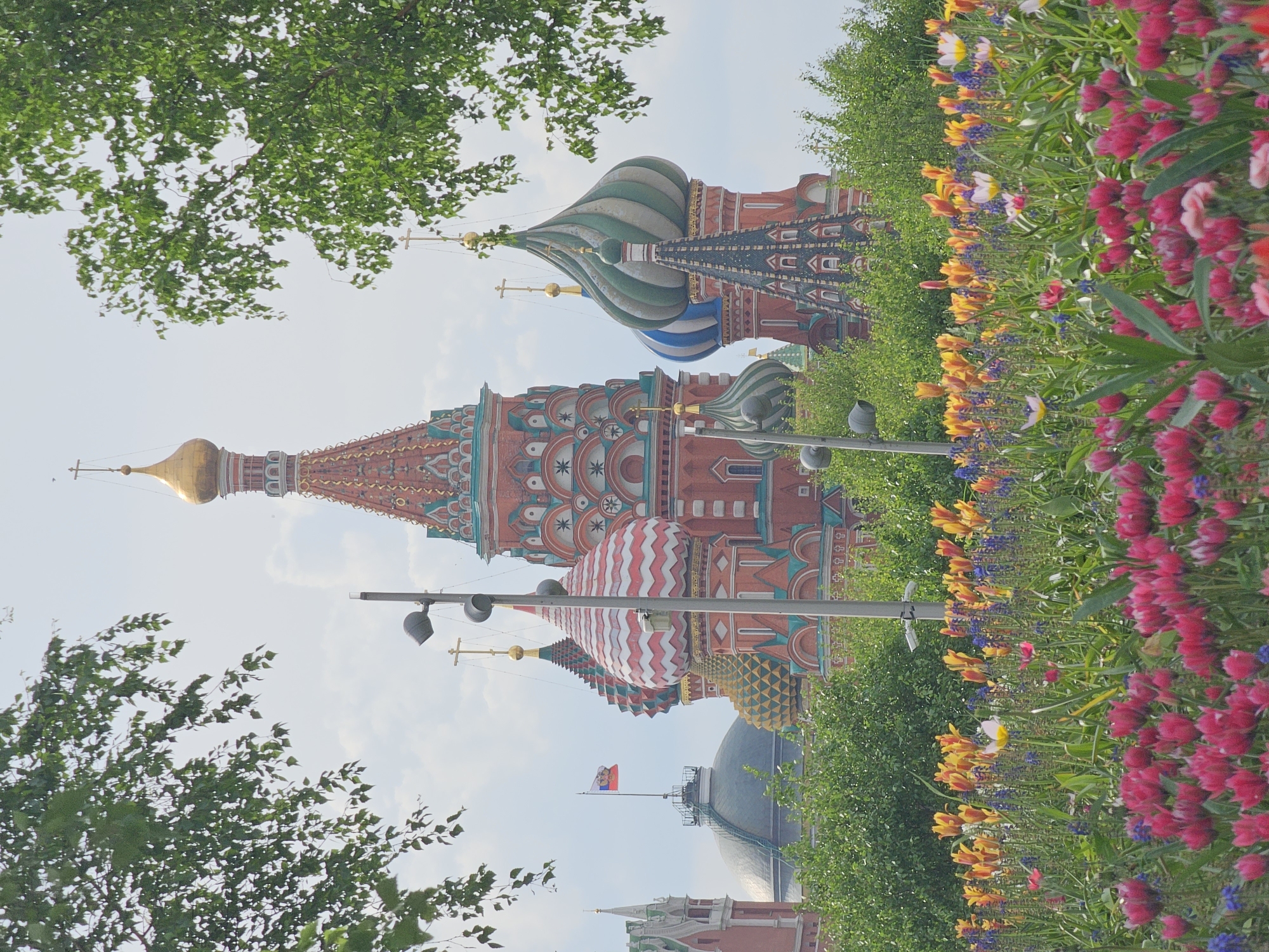The image shows a view of St. Basil's Cathedral in Moscow, Russia, framed by trees and a vibrant flowerbed. The cathedral's ornate domes are visible, featuring a variety of colors and patterns, including red, white, green, and blue. A Russian flag flies atop a nearby structure. The flowerbed contains a mix of red, yellow, and purple flowers. Streetlights are interspersed among the flowers and trees. The sky is partly cloudy.