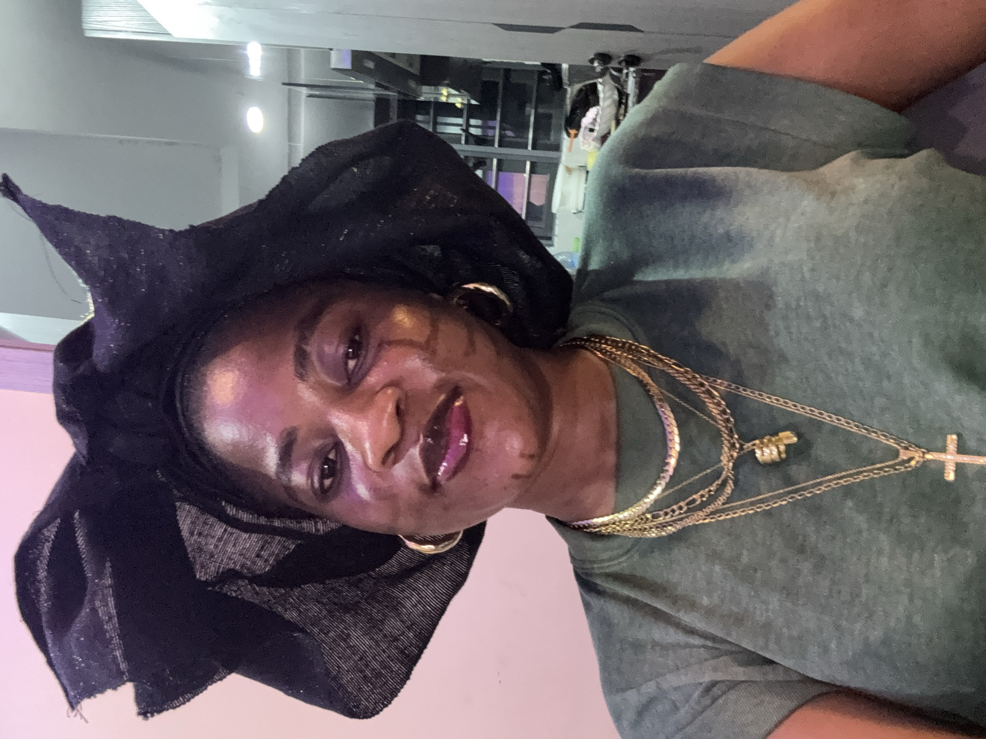 This is a medium shot, portrait-oriented photograph of a woman. She is looking towards the camera with a slight smile and her head tilted. The woman is wearing a black headwrap with a pointed design at the top, gold hoop earrings, and multiple gold necklaces, one of which has a cross pendant. Her attire includes a green crew neck shirt. The lighting suggests an indoor setting, possibly with overhead lights reflecting on her face and the background. The background appears to be a room with some shelving units and possibly merchandise visible, hinting at a retail or display area. There are no visible text elements in the image. The overall mood seems relaxed and confident. The photograph is rotated 90 degrees clockwise.
