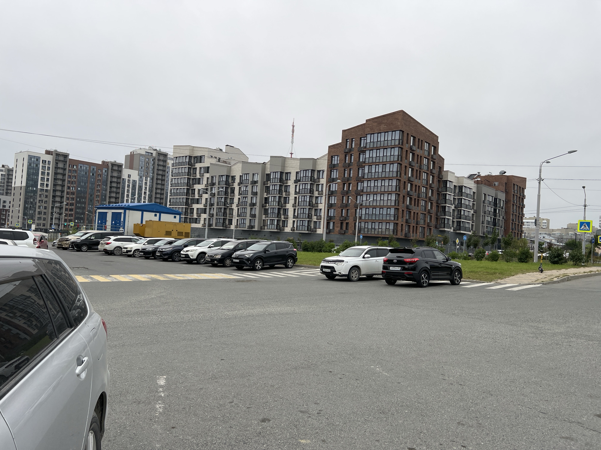 The image shows a street scene in Yuzhno-Sakhalinsk, Russia, featuring several modern apartment buildings and a parking lot filled with cars. The apartment buildings are tall, with varied architectural styles, including brick facades and large windows. The sky is overcast, suggesting a cloudy day. In the foreground, a silver SUV is parked on the left side of the frame. Behind it, a line of various cars is parked in a designated lot. A pedestrian crossing is visible on the right side of the image. The environment appears to be a newly developed urban area. There are no visible people actively engaged in any specific actions.