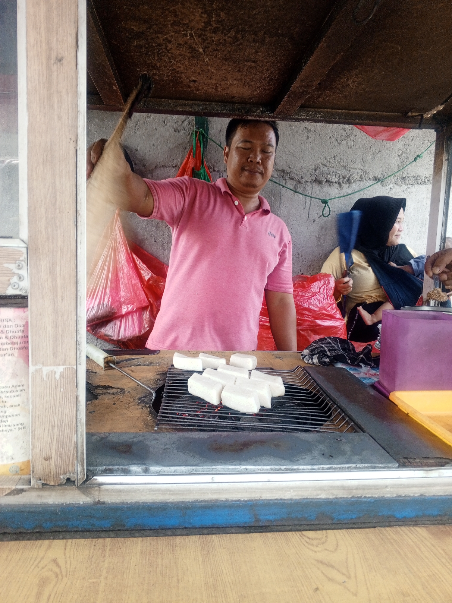 This is a medium shot, eye-level photograph taken inside a food stall in Depok, Indonesia. A man, appearing to be in his 30s or 40s, wearing a pink polo shirt, stands behind a charcoal grill. He is holding a long-handled tool, possibly for flipping food, and looking towards the viewer. On the grill, several rectangular pieces of white food, likely tofu or tempeh, are being cooked over glowing red coals. Behind the man, a woman wearing a black hijab is visible, holding a baby who is partially visible. Various items are present in the stall, including red plastic bags, a blue plastic fan, and containers. The stall appears to be made of wood and metal, with a textured wall in the background. The overall lighting suggests it is daytime.