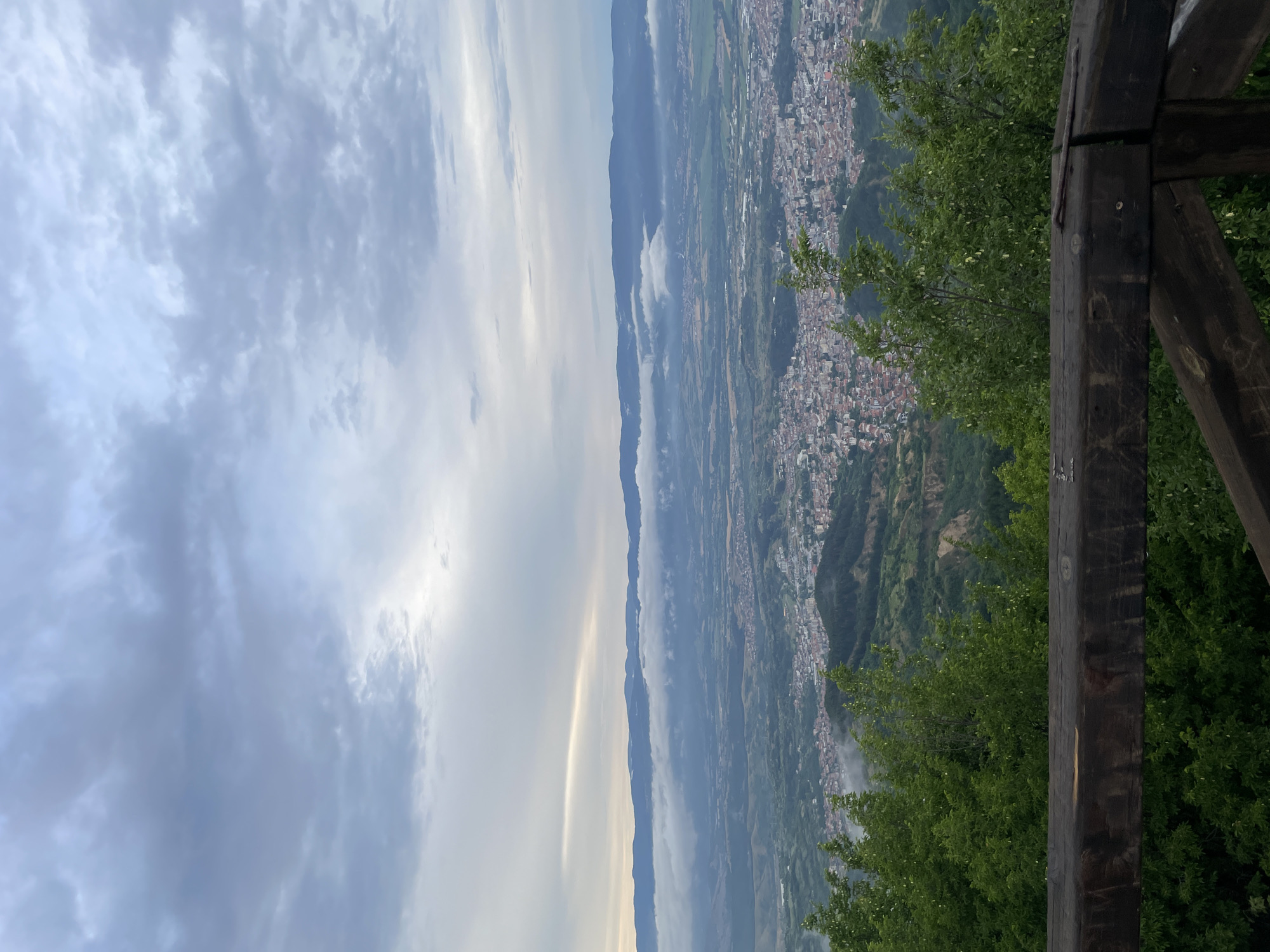 A high-angle, full shot captures a view of Delchevo, Bulgaria, from a lookout point. The foreground is dominated by a dark, weathered wooden railing. Beyond the railing, lush green trees form a dense canopy, their leaves slightly blurred. In the middle ground, the city of Delchevo sprawls across a valley, its buildings characterized by red-tiled roofs. The city is nestled among rolling green hills. In the background, a mountain range stretches across the horizon, partially obscured by a band of soft, white clouds that hug the lower slopes. The sky above is overcast with layers of gray and white clouds, with a faint strip of lighter sky visible near the horizon.