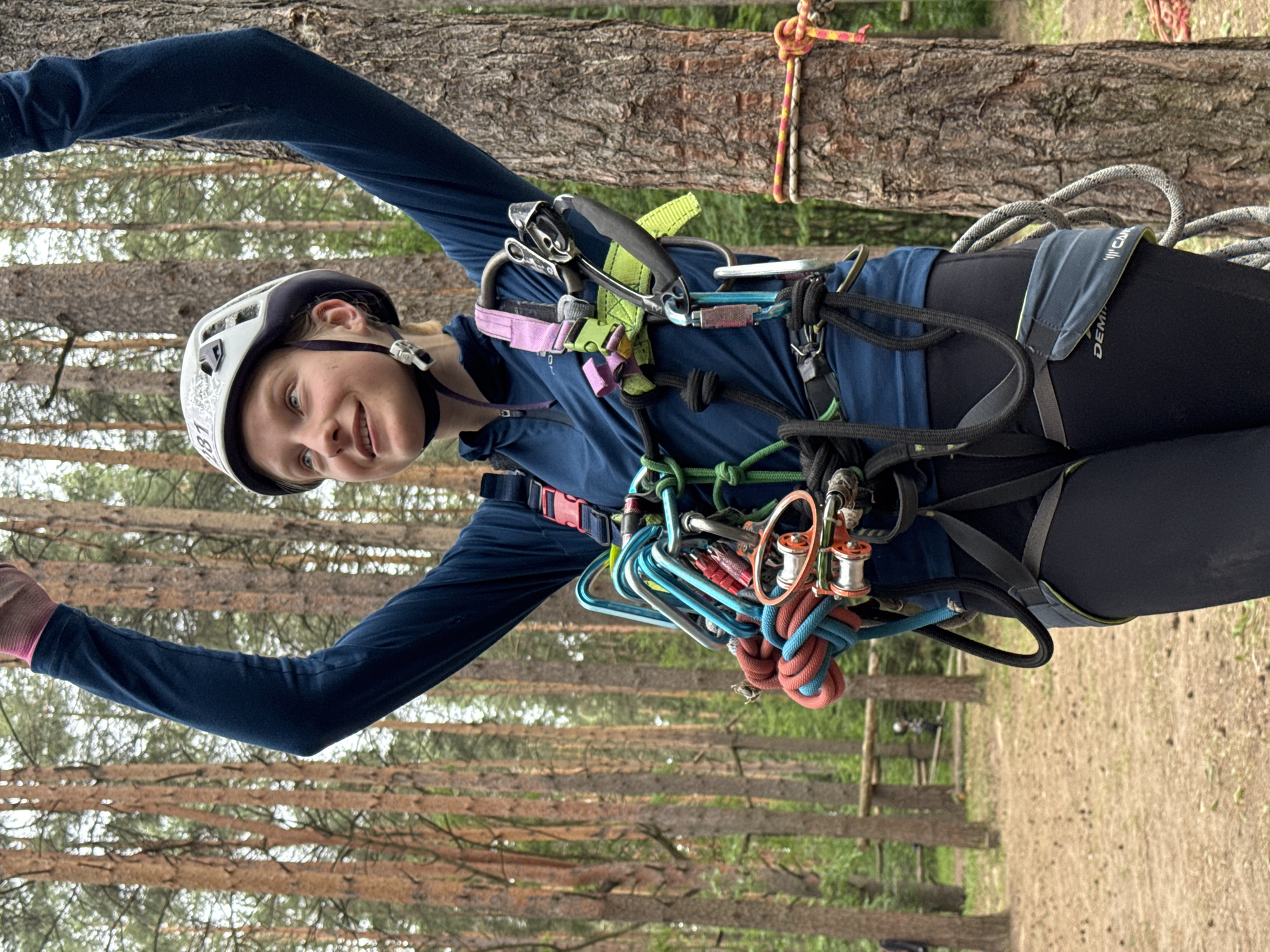 A young Caucasian girl, likely in her early teens, is standing outdoors in a forest, possibly at an adventure park in Chaikovsky, Russia. She is wearing a blue long-sleeved shirt, black leggings, a white climbing helmet, and a full climbing harness with various carabiners, ropes, and ascenders attached. Her arms are raised above her head in a gesture of triumph or excitement, and she is smiling. The background consists of tall pine trees, with a sandy, wooded ground cover. There are wooden poles and ropes visible in the middle ground, suggesting an obstacle course or climbing area. The lighting indicates it is daytime, possibly overcast or early morning/late afternoon, given the soft light and muted colors. The girl's attire and equipment suggest she has just completed or is about to begin a climbing or aerial adventure activity.