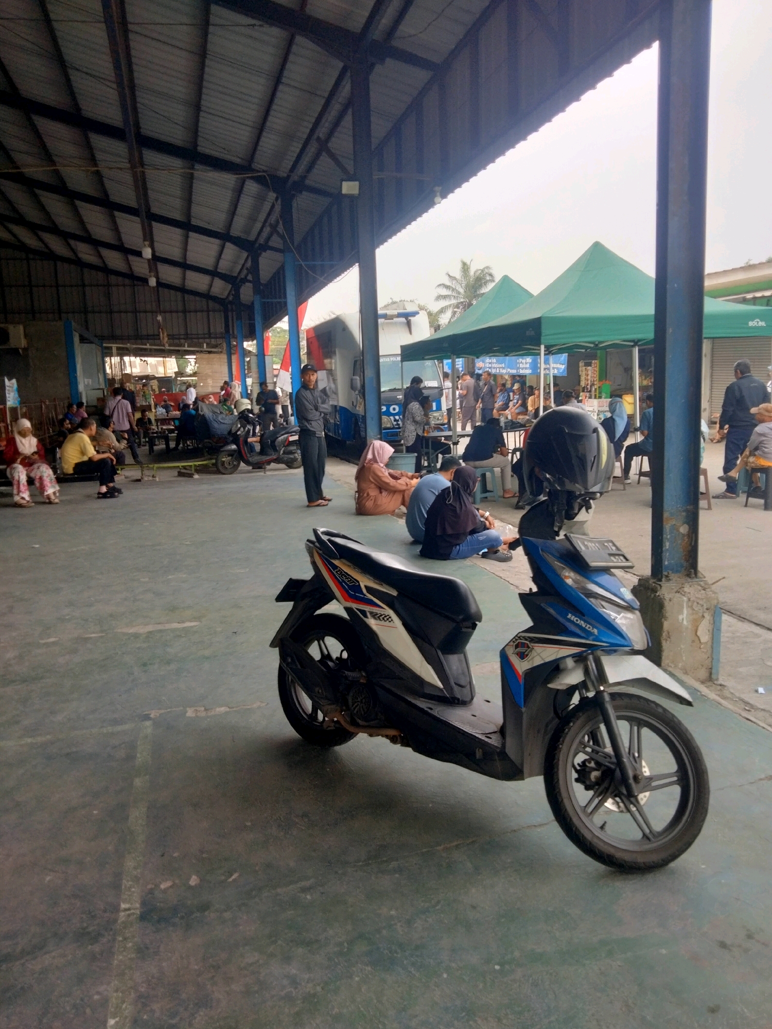 The image depicts an outdoor scene under a covered structure, likely a marketplace or a waiting area, in Sukaasih, Indonesia. A blue and white motorcycle is parked in the foreground, facing left. Behind it, several people are seated or standing. Some are gathered under green tents in the middle ground, where food or merchandise might be displayed. In the background, more people are visible, and a large vehicle, possibly a bus, can be seen. The ground is paved concrete. The overall atmosphere appears to be casual, with people engaged in various activities, including waiting and socializing. The lighting suggests it is daytime.