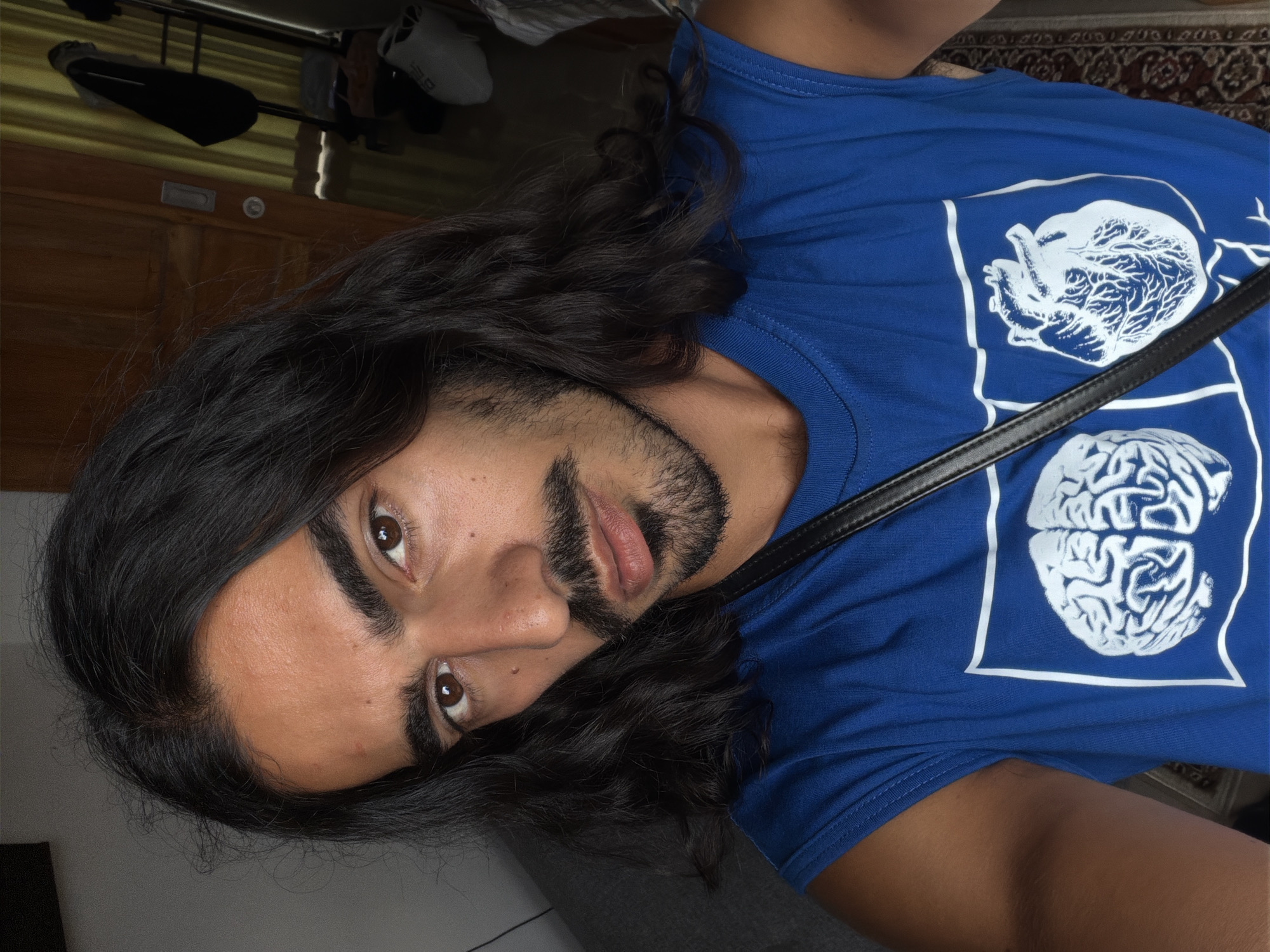 A medium-shot photograph, oriented vertically, shows a person with dark hair and a beard, wearing a blue t-shirt with a white graphic of a heart and brain. The person's gaze is directed slightly downward, their head tilted towards the left. The background includes a door and some items on a shelf. The location of the photograph is North Kuta, Indonesia.