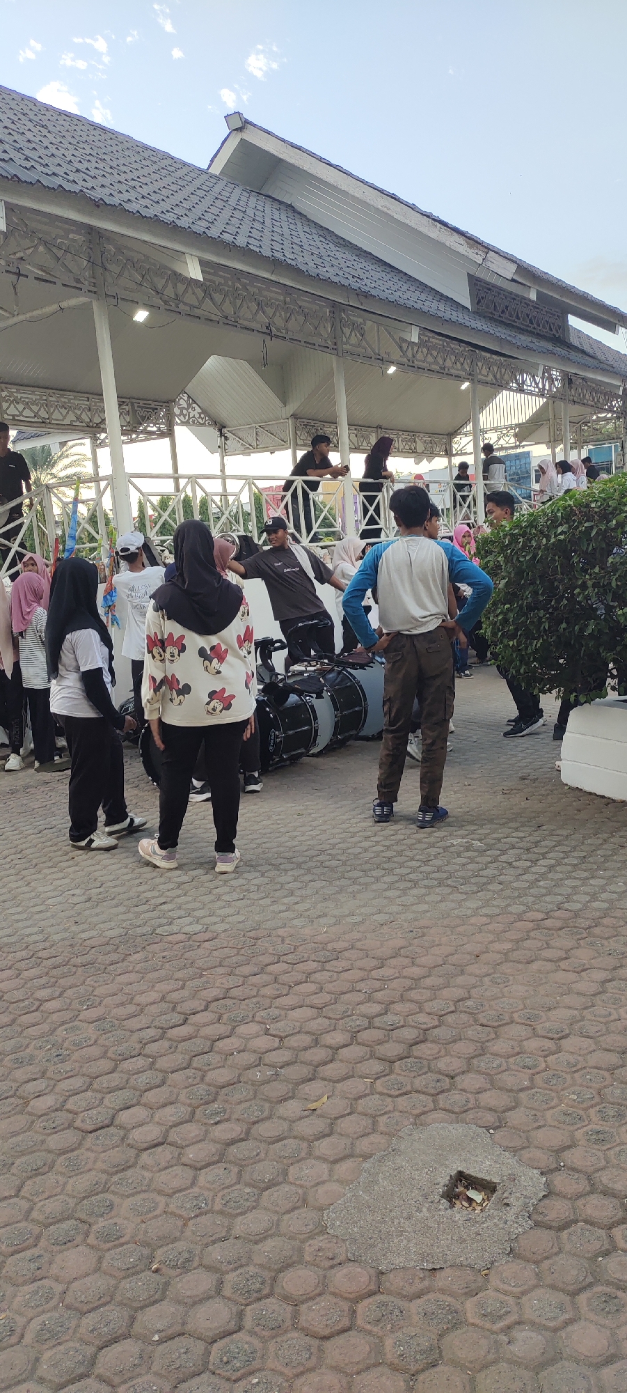 In Lhokseumawe, Indonesia, a group of young people are gathered around large drums, possibly for a marching band or performance. They are positioned on a paved outdoor area with hexagonal-shaped bricks. Overhead, a white, covered pavilion with a dark, tiled roof provides shade. Several individuals are wearing headscarves, indicating a cultural context. One person in the foreground is wearing a light-colored shirt with a Minnie Mouse print. Another person in a blue shirt and brown pants stands with hands on hips, looking away from the camera. The sky above is a pale blue with scattered clouds. The overall scene suggests a warm, daytime gathering.