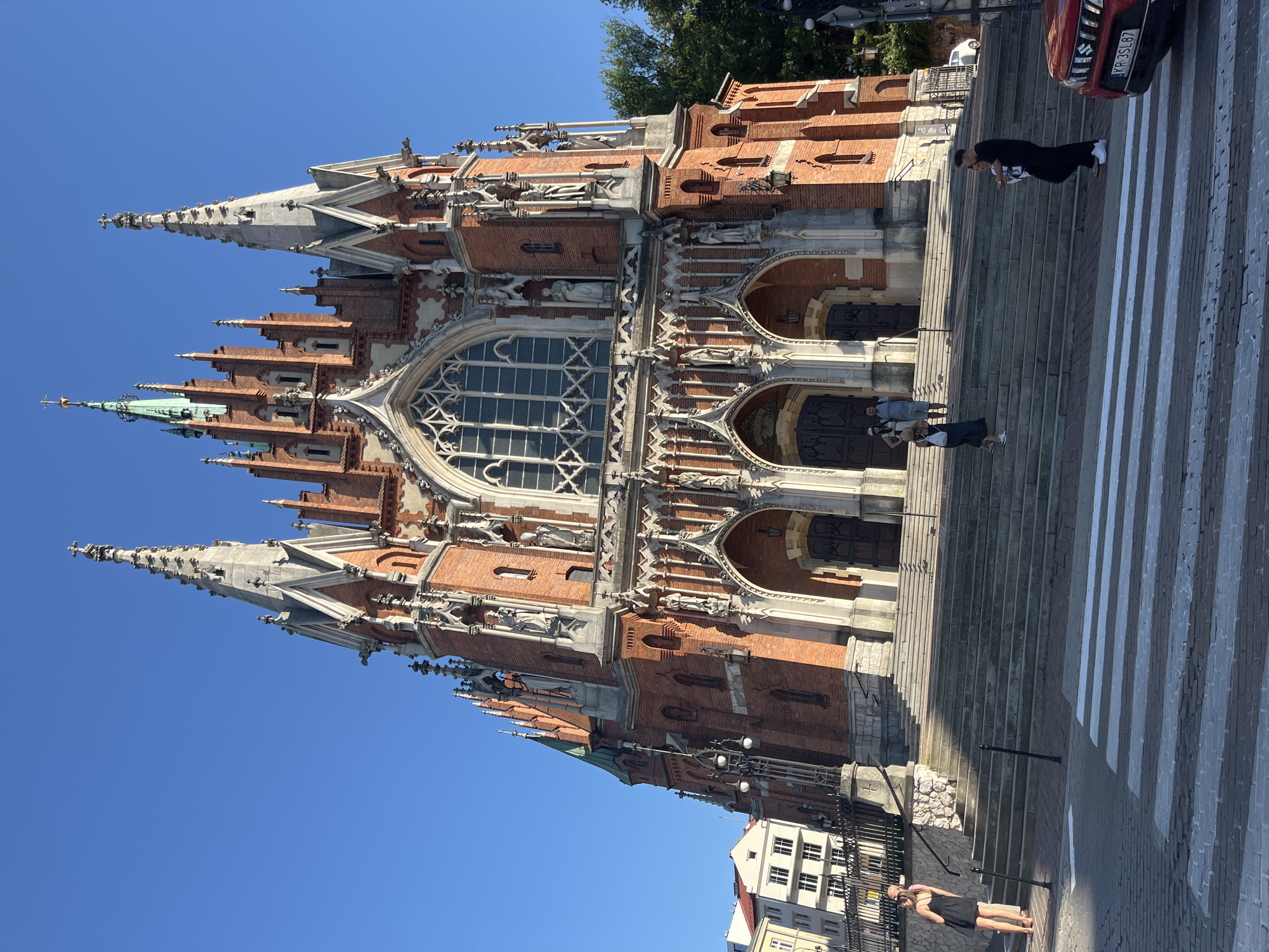 The image shows the Church of Saints Peter and Paul in Krakow, Poland, on a clear, sunny day. The grand brick facade of the church, with its ornate spires and Gothic architecture, dominates the upper portion of the frame. A wide, stone staircase leads up to the church entrance, where several people are visible. In the foreground, a pedestrian crossing with white stripes is marked on the cobblestone street. A red car is parked on the right side of the street, with its license plate visible. A few trees are visible on the right side of the image, providing a contrast to the architectural elements. The sky is a vibrant blue.