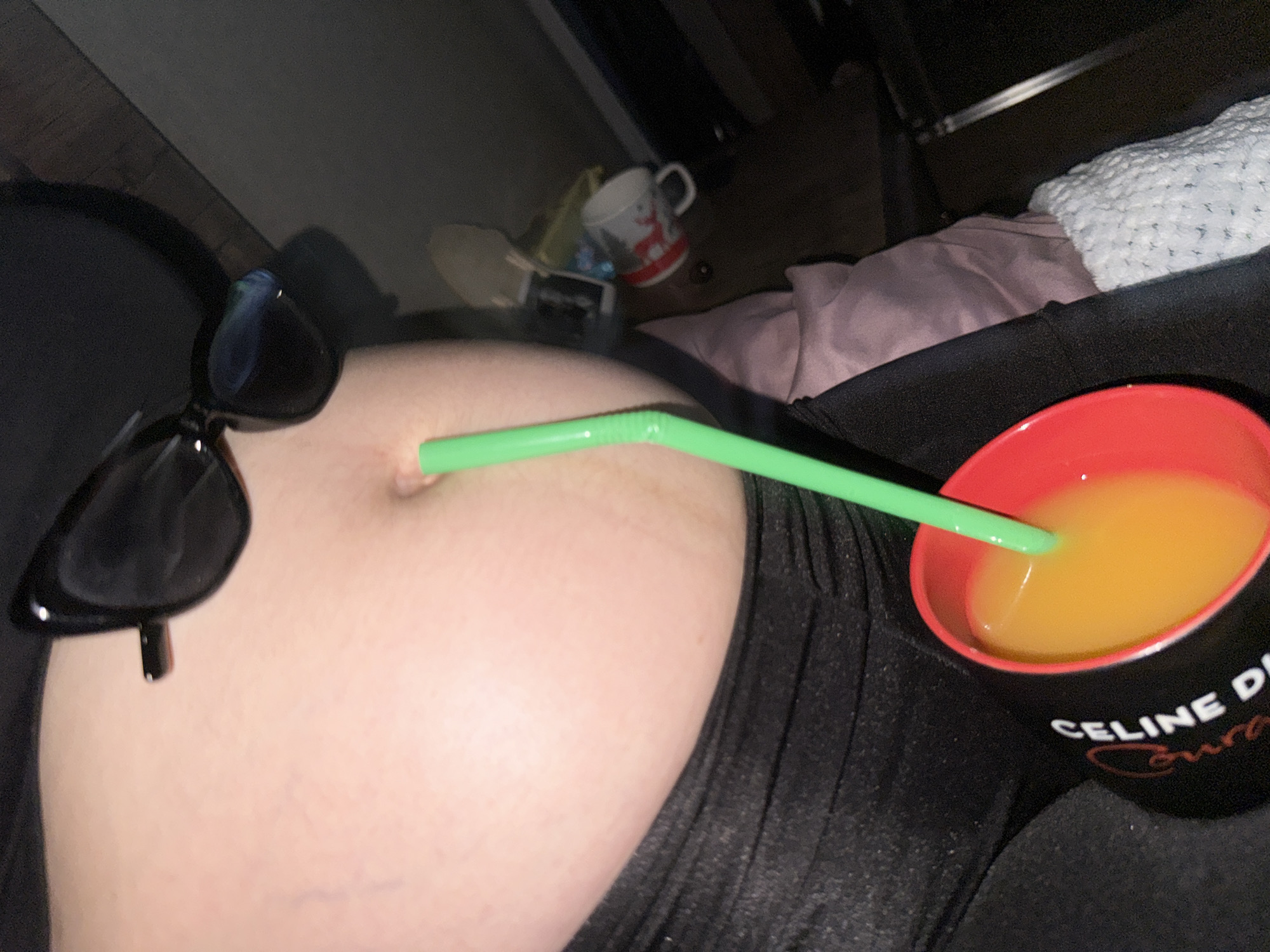 The image is a close-up shot of a person's midriff and belly, with a green straw inserted into their belly button. The person is wearing black leggings and a black top. They are also wearing black sunglasses perched on their stomach. A red cup filled with orange liquid and a green straw is held in their hand. In the background, there are some objects on a shelf, including a mug. The scene appears to be indoors, possibly in a room in Tbilisi, Georgia. The lighting is dim, suggesting it might be nighttime or a poorly lit area.