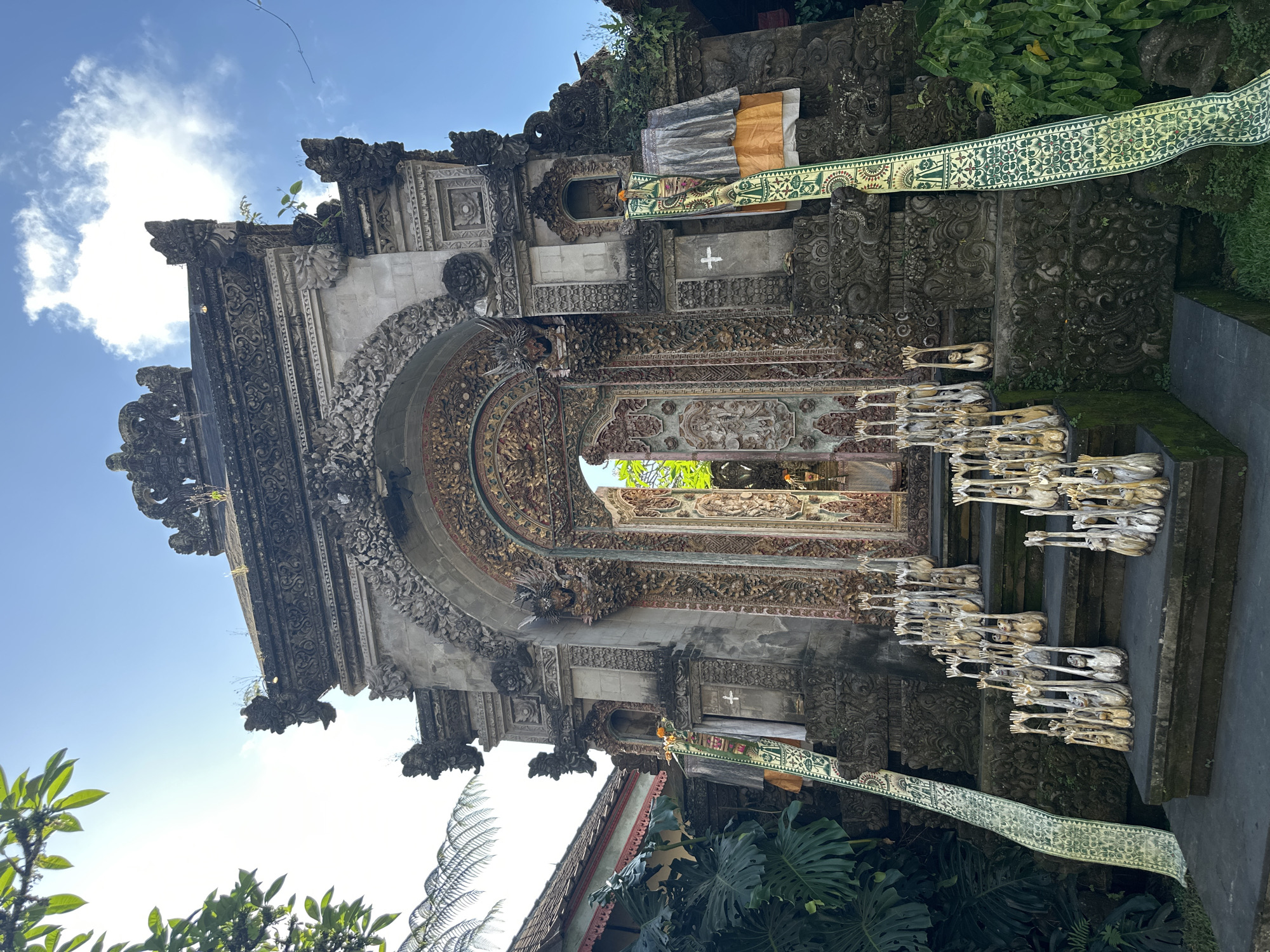 This image depicts a detailed, ornate temple entrance in the Ubud District of Indonesia. The entrance is a grand structure with intricate stone carvings and a large, arched gateway leading into the temple. The gateway is adorned with detailed decorative panels and features two intricately carved doors, which are partially open.
The steps leading up to the temple entrance are lined with numerous small, wooden figures, each with its hands raised. A long, patterned green and white fabric banner hangs vertically along the left side of the steps, and a similar banner is visible on the right side of the entrance. The temple is surrounded by lush green foliage, with large tropical leaves and trees visible on the left and right sides of the frame. The sky above is a clear blue with a few scattered white clouds. The architecture and decorations suggest a Balinese Hindu temple.