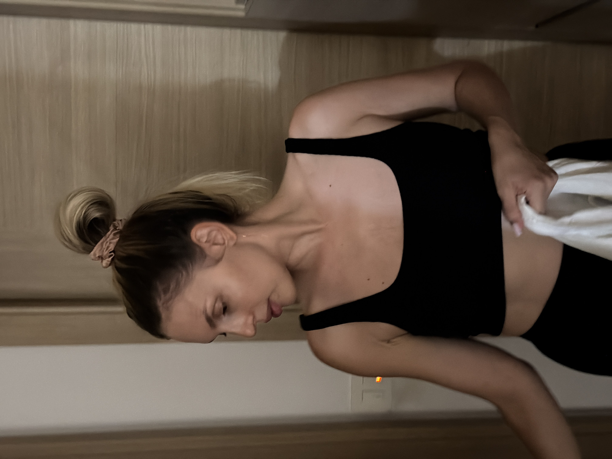 A medium shot captures a woman with her hair in a bun, wearing a black crop top. She is looking down and to her left, with her left arm bent and holding a white cloth. The background consists of a neutral-colored wall and a wooden door. An illuminated light switch is visible on the left side of the frame. The setting appears to be an indoor space, possibly a room in Budva, Montenegro, given the context provided.