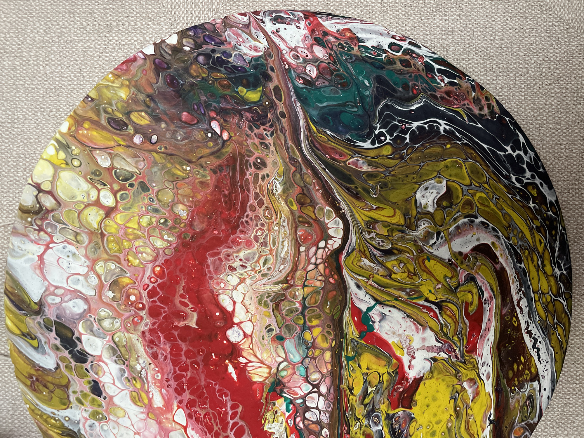 The image shows a close-up, overhead view of a circular abstract painting. The painting is a fluid art piece, characterized by swirling patterns and cells of various colors. Dominant colors include vibrant red, yellow, green, black, white, and shades of brown and purple. The paint is applied in a way that creates organic, interconnected shapes and textures, resembling cellular structures or flowing liquids. The painting is placed on a neutral-colored, textured surface, likely a canvas or fabric backdrop, which provides a subtle contrast to the artwork. There is no discernible location in the city of Zheleznogorsk, Russia depicted in this image; it is solely a depiction of an abstract painting.