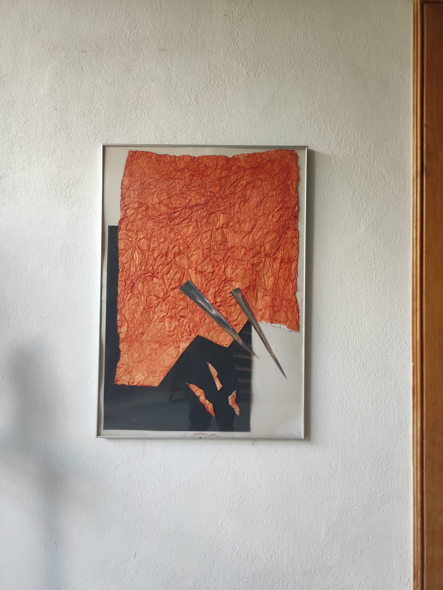 A framed abstract artwork is displayed on a textured white wall, to the right of a wooden doorframe. The artwork features a large piece of crumpled, vibrant orange paper as the dominant element. Below the orange paper, there are abstract shapes in black and white. Two metallic, pointed objects, resembling sharpened metallic blades or stylized wings, are placed diagonally across the artwork, overlapping both the orange paper and the black shapes. The metallic objects appear to be positioned in front of the paper, adding a three-dimensional aspect to the collage. The overall impression is one of sharp contrast and dynamic composition, with the textured paper and metallic elements creating visual interest. The lighting suggests it is daytime, with soft illumination on the wall and artwork, creating subtle shadows. The setting appears to be an interior space, likely a home or gallery. There is no visible text in the image.