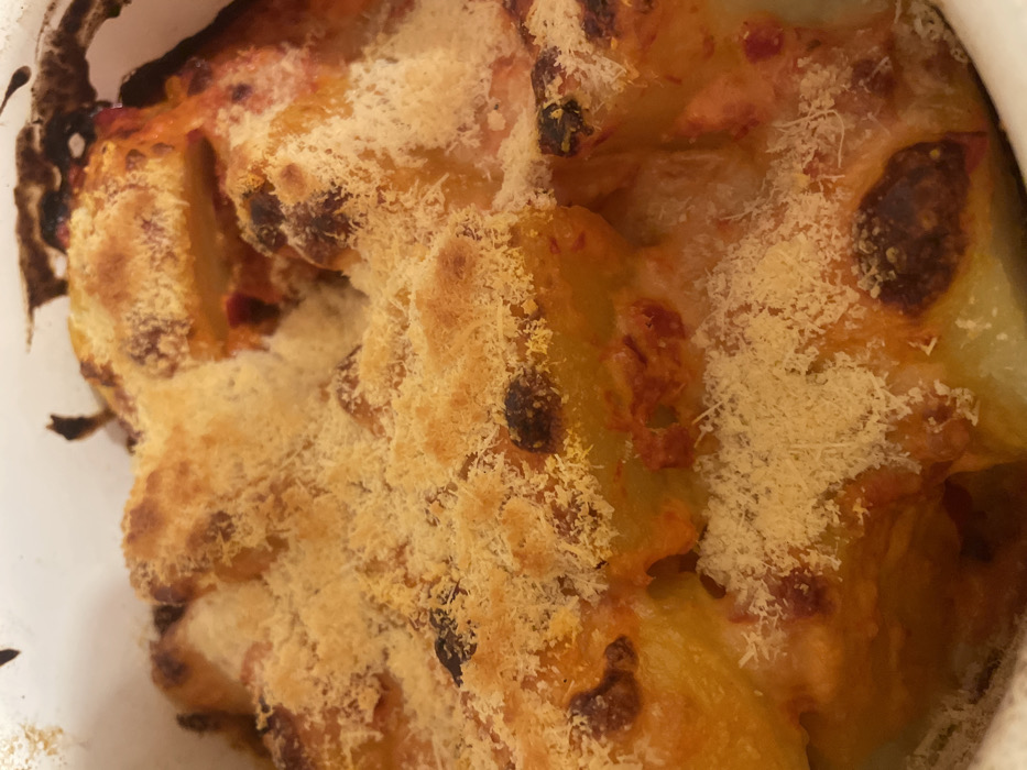 A close-up, overhead view displays a portion of a baked dish, observed in the city of Palež, Montenegro, contained within a white, possibly ceramic, oven-safe vessel. The primary subject is a food item characterized by a golden-brown, granular crust, consistent with baked cheese or breadcrumbs. This crust exhibits varying degrees of thermal browning, ranging from light gold to darker, almost burnt patches, with numerous small, dark specks interspersed throughout. Beneath the crust, larger, irregularly shaped, light-colored components, suggestive of par-cooked potatoes or pasta, are visible. These components are partially submerged in and coated with a reddish-orange, viscous sauce, indicative of a tomato or pepper base. The dish exhibits signs of recent baking, including bubbling textures and caramelized edges on some of the underlying food items and sauce. No individuals, specific actions, or direct interactions are depicted. The immediate environment captured is confined to the food and the interior edge of the serving vessel.