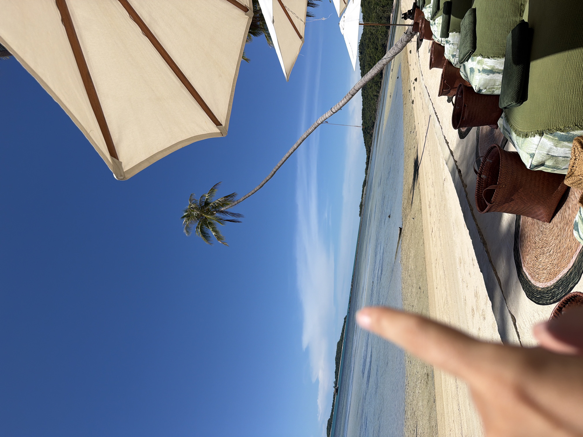 The image depicts a tropical beach scene in Baan Thong Krut, Thailand. The foreground features a blurred human finger pointing towards the ocean. The middle ground shows a sandy beach lined with comfortable-looking green lounge chairs, several woven baskets, and a circular straw mat. Beyond the beach, the calm, shallow ocean stretches towards a distant, tree-covered island under a clear blue sky with a few wispy clouds. A distinctive palm tree arches dramatically over the beach, leaning towards the water. Several white and off-white umbrellas are positioned to provide shade for the lounge area. The overall impression is one of a serene and idyllic beach resort setting.