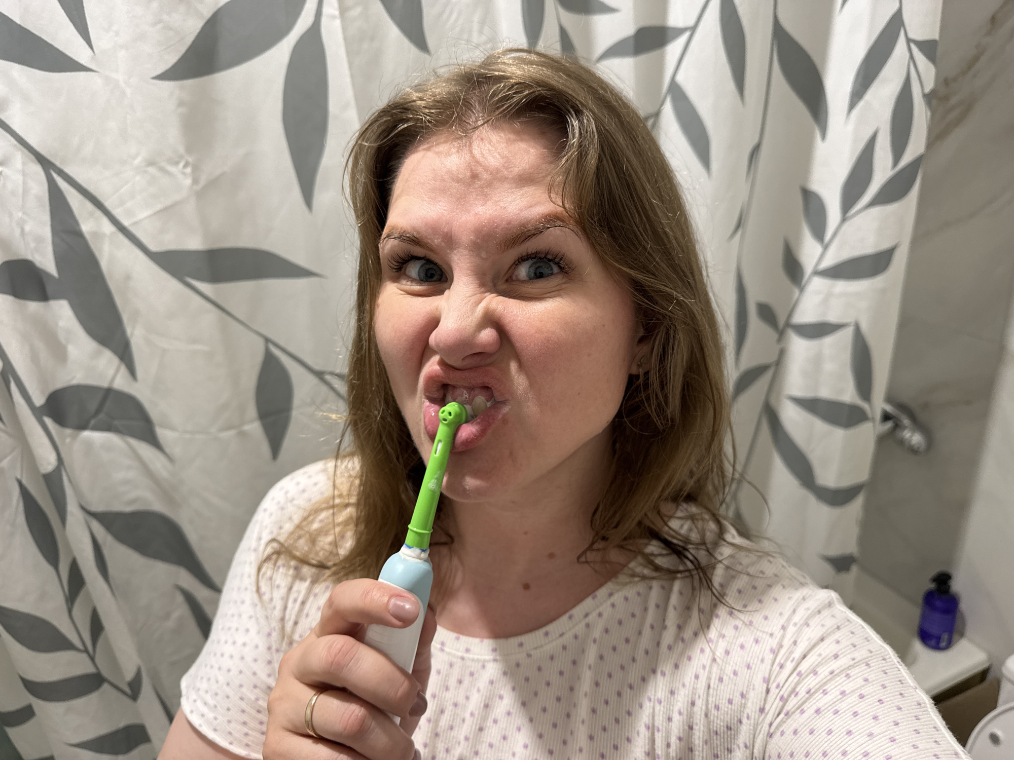 A woman is taking a selfie in a bathroom. She is in front of a shower curtain with a gray and white leaf pattern, holding an electric toothbrush in her mouth. The toothbrush is light blue and green with a white handle. She is making a funny face, squinting and wrinkling her nose. She is wearing a white t-shirt with light purple polka dots. Behind her to the right is a shelf with a blue bottle on it. Based on the availability of shower curtain and toiletries, it appears this photograph was taken in a private residence or a hotel room. There is no evidence of location in the city of Gornyi (Dvurechenskoe S/P), Russia.