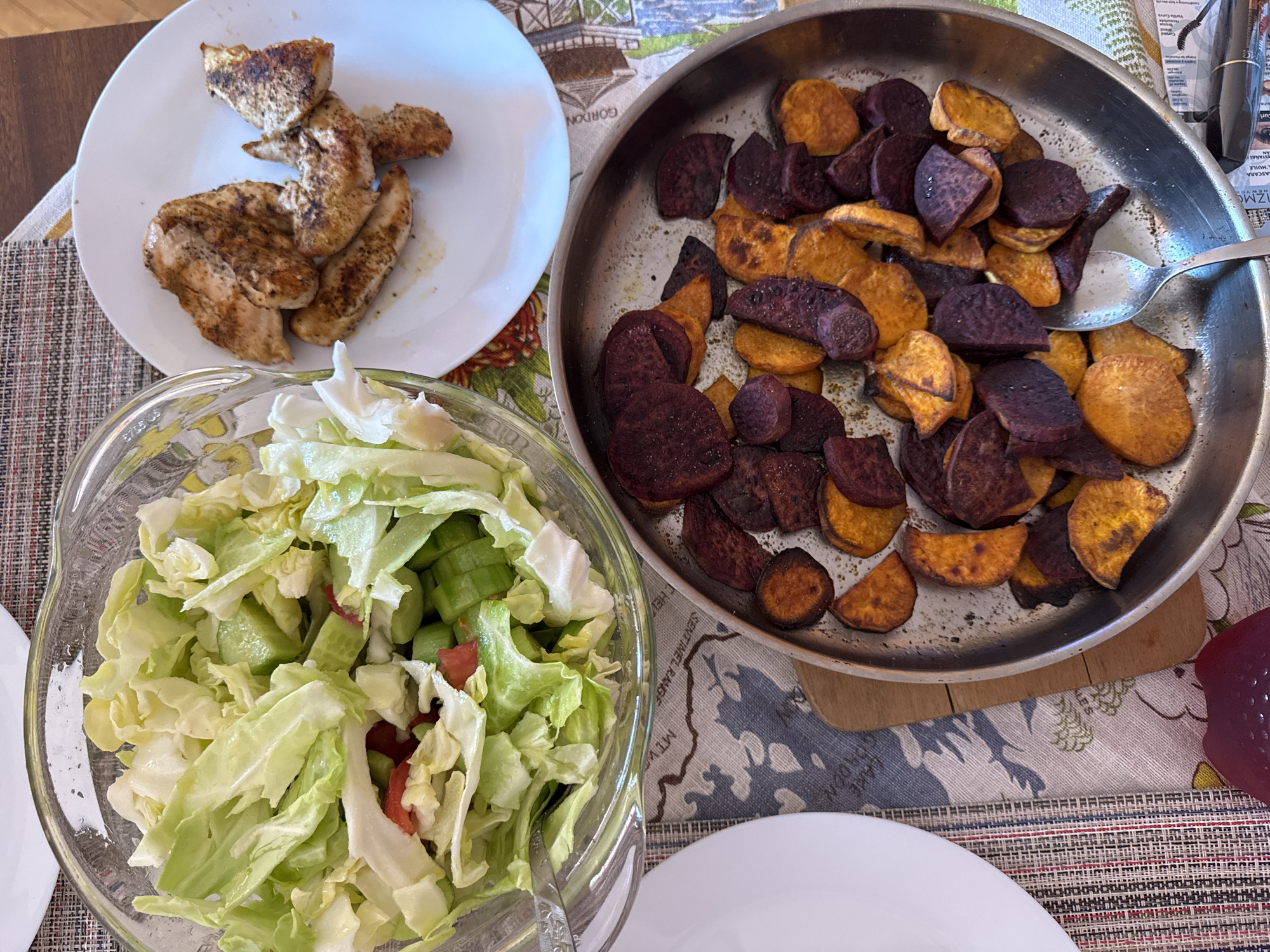 The image shows a table setting with a meal consisting of grilled chicken on a white plate, a salad in a clear glass bowl, and a pan of roasted purple and orange sweet potato slices. A spoon is present in the pan. The table has a patterned tablecloth. The location in Burgas, Bulgaria is not determinable from the image.