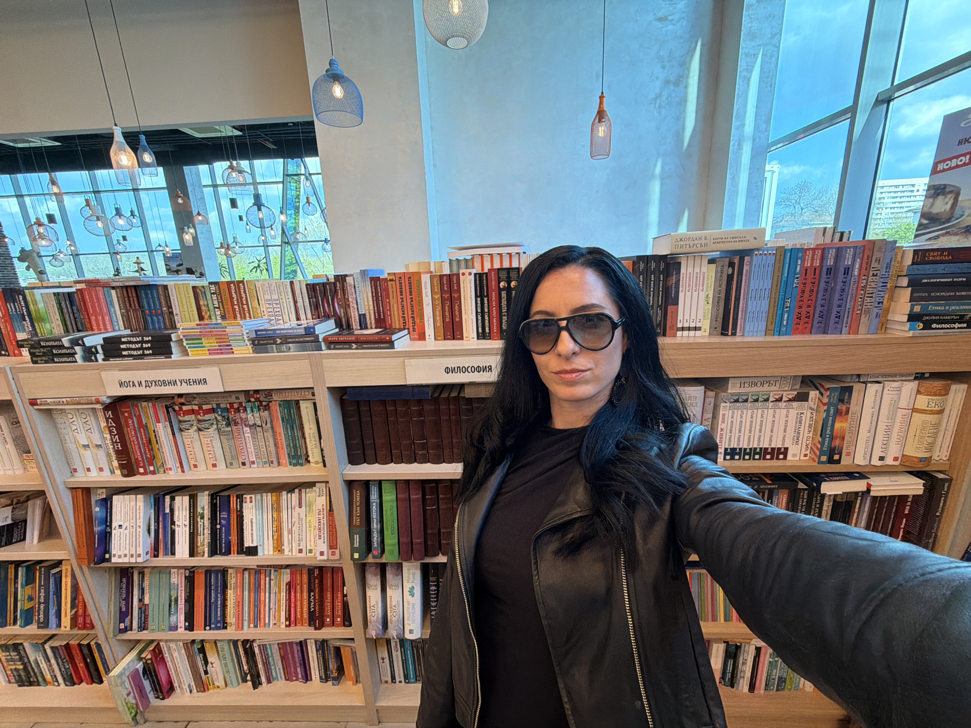 A woman in sunglasses and a black leather jacket is taking a selfie inside a bookstore. She is positioned in front of bookshelves filled with books, some of which have titles in Bulgarian. The shelves are labeled with categories such as "ЙОГА И ДУХОВНИ УЧЕНИЯ" (Yoga and Spiritual Teachings) and "ФИЛОСОФИЯ" (Philosophy). The location is likely a bookstore in the city of Pleven, Bulgaria. Visible through windows are buildings and the sky. Hanging pendant lights are seen from the ceiling.