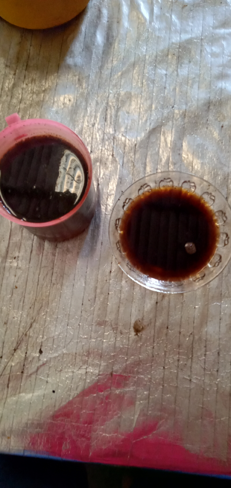 The image shows two containers, a pink mug and a small glass bowl, both filled with a dark liquid, likely coffee or tea, resting on a textured, slightly stained surface. The background is indistinct, suggesting an indoor setting. The picture was taken in Sumput, Indonesia.