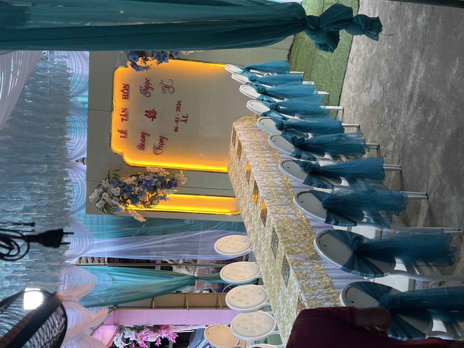 This image captures a beautifully decorated setting for what appears to be a wedding or engagement ceremony in Bình An, Vietnam.
The central focus is a long, rectangular table draped with a white and gold patterned tablecloth. On one side of the table are several white circular stools, while the opposite side features white chairs adorned with elegant teal fabric sashes tied in bows, facing the table. Beyond these chairs, the flooring transitions from concrete to artificial green turf.
At the head of the table stands a prominent, illuminated white sign. This sign prominently displays Vietnamese text: "LỄ TÂN HÔN," which translates to "Wedding Ceremony" or "Engagement Ceremony." Below this, the names "Quang Trọng" and "Ngọc" are visible, likely the names of the couple. Between their names is the traditional Chinese "Double Happiness" symbol, often used in weddings. The date "06-03-2026" is also inscribed, followed by "AL," possibly referring to the solar calendar date.
Floral arrangements featuring blue, white, and purple flowers decorate the base of the illuminated sign. The surrounding area is draped with white sheer fabric and teal-colored cloth, creating a festive and elegant ambiance. A person's arm and shoulder, wearing a dark red shirt, are partially visible in the bottom right, suggesting guests are present. An electrical plug and what might be part of a fan or light fixture are visible in the bottom left foreground. The overall impression is one of celebration and joyous occasion, likely an important family event given the formal decorations and signage.
