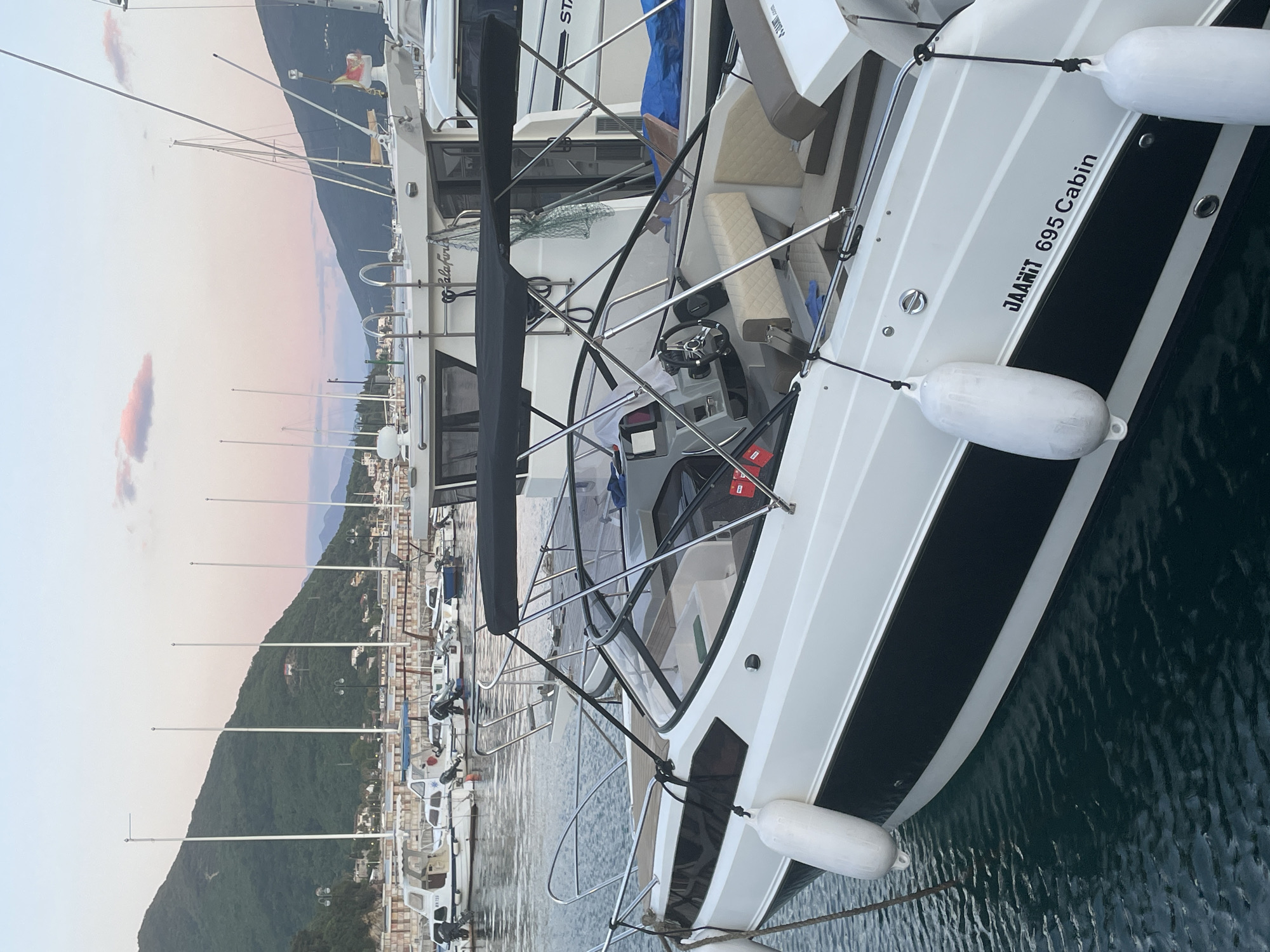 This media file captures a serene evening or morning scene in a marina in Meljine, Montenegro. The primary subject is a white motor yacht, prominently docked in the foreground, with its port side facing the viewer. The boat features a distinctive black stripe along its hull, contrasted with the white upper body, and is protected by two white fenders. Inside the cabin cruiser, a steering wheel, helm console, and light beige upholstered seating are visible, suggesting a comfortable interior.
In the background, a large, dark green, tree-covered mountain range dominates the landscape, receding into the distance. The sky above is painted with soft hues of light blue, fading into delicate pinks and oranges, indicative of either sunrise or sunset. The water in the marina is calm, reflecting the ambient light and showing gentle ripples.
Numerous other boats, including smaller fishing or leisure vessels and those with tall masts, are docked along the marina, stretching towards the base of the mountains. Some distant buildings with faint lights are visible along the shore. A flag, possibly Montenegrin with red and yellow/gold elements, is seen flying from a boat further back.
Visible text on the main boat clearly reads "JAARIT 695 Cabin". On a boat directly behind it, partially obscured text appears to be "PalaFiri" or "PalaFiori". Another boat further down the dock has the registration "MV 7733". Partially visible text "STARC" or similar can be seen on the bow of a boat in the upper left of the main vessel. The overall atmosphere is calm and peaceful, suggesting good weather conditions.