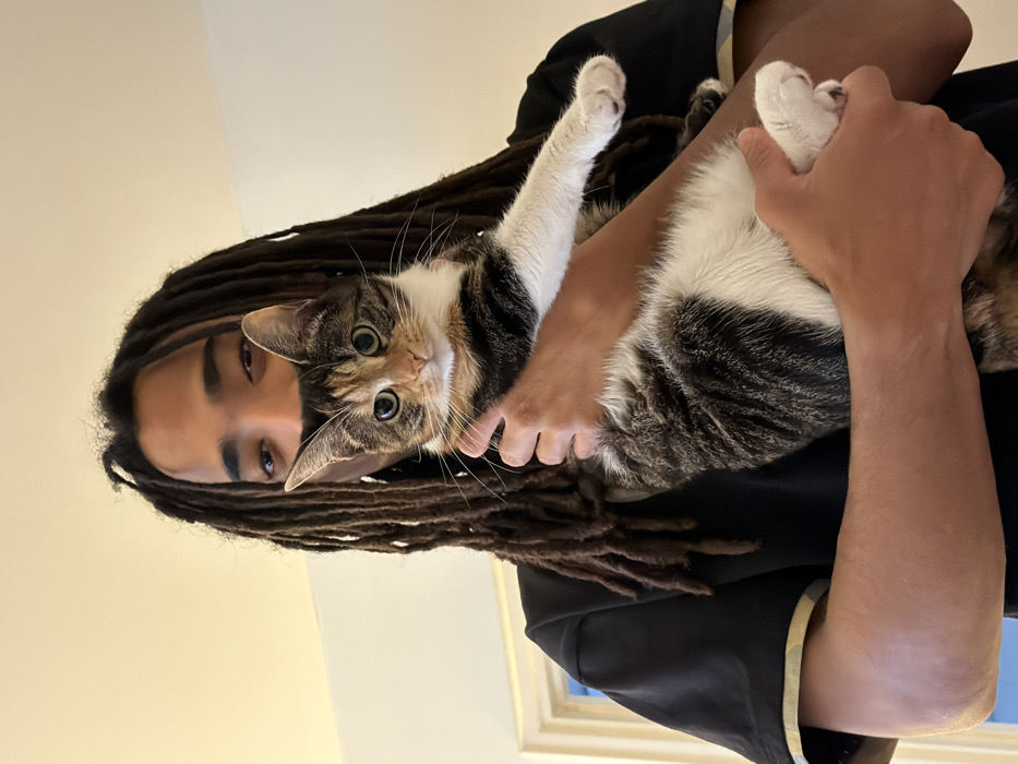 A person with dreadlocks is holding a cat. The cat, which is a mix of white, tabby, and brown, is looking directly at the camera. The person is wearing a black shirt with a yellow and white trim. The background is a light beige wall and a window frame is visible on the right side of the image.