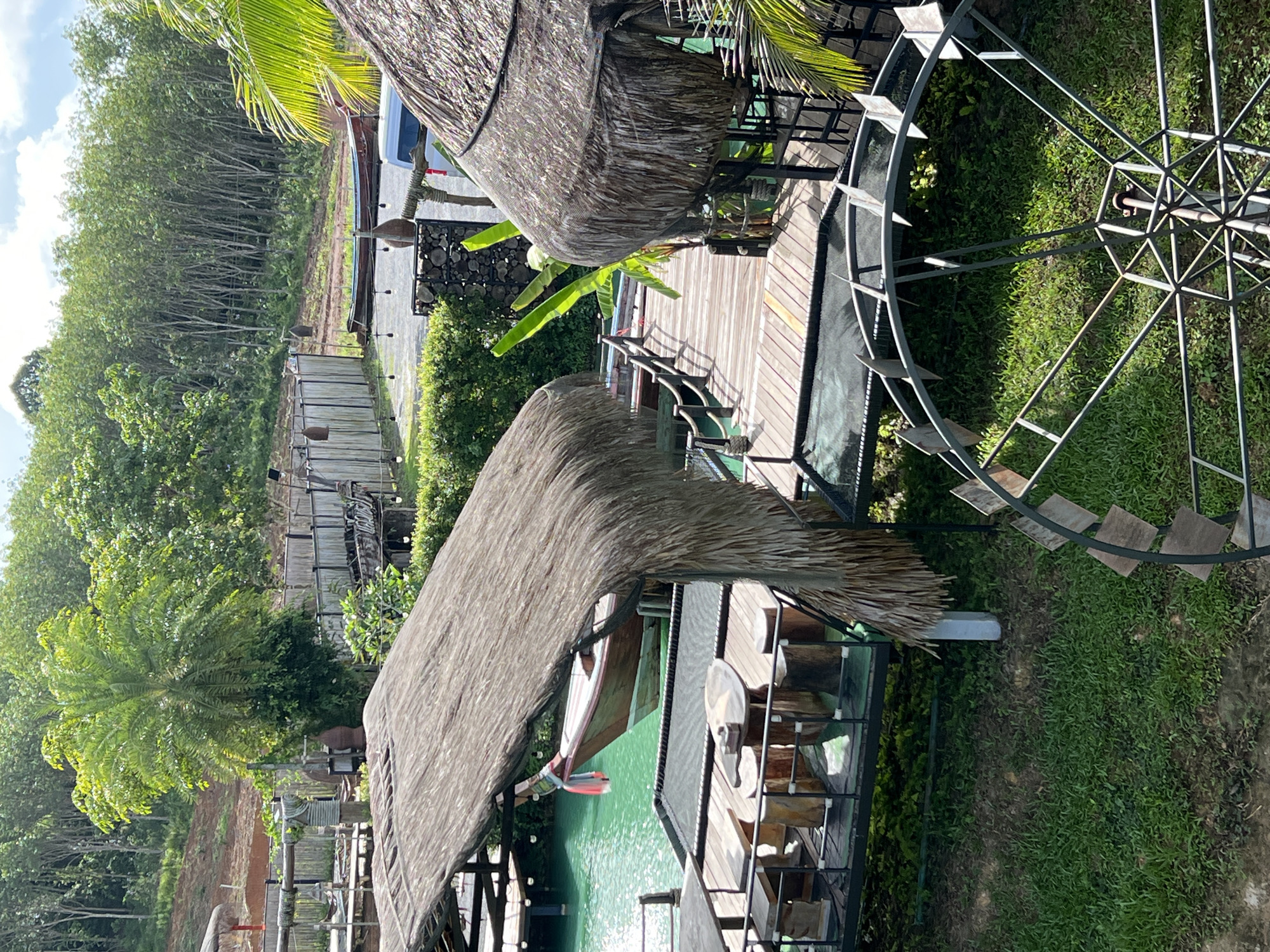 The image shows an outdoor seating area with thatched-roof structures and wooden decks overlooking a body of turquoise water. In the foreground, a metal water wheel structure is visible on the grass. A wooden boat is moored to the water's edge, and a palm tree and other lush vegetation are present in the background, with a densely wooded hillside beyond. The scene appears to be a tranquil resort or restaurant setting.