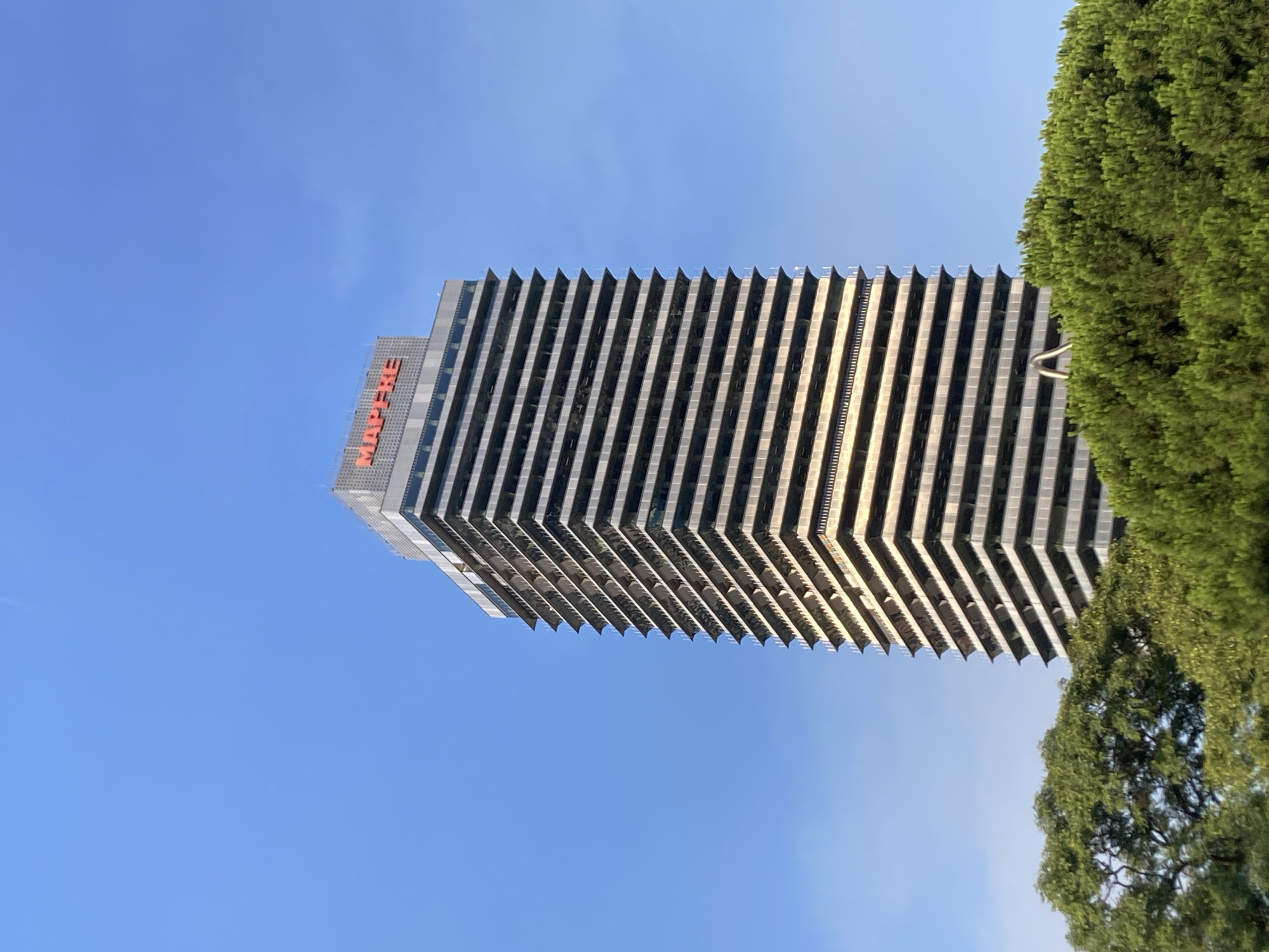 The image shows the Mapfre Tower, a skyscraper located in the Port Olímpic area of Barcelona, Spain. The tower, with its distinctive red "MAPFRE" signage at the top, is a prominent feature of the city's skyline. It is surrounded by lush green trees, with the bright blue sky serving as the backdrop. The building's facade is characterized by horizontal bands of windows and balconies, giving it a layered appearance. The perspective suggests the photo was taken from below, looking up at the tower. No people or vehicles are visible in the frame. The lighting indicates it is daytime, likely with clear weather.
