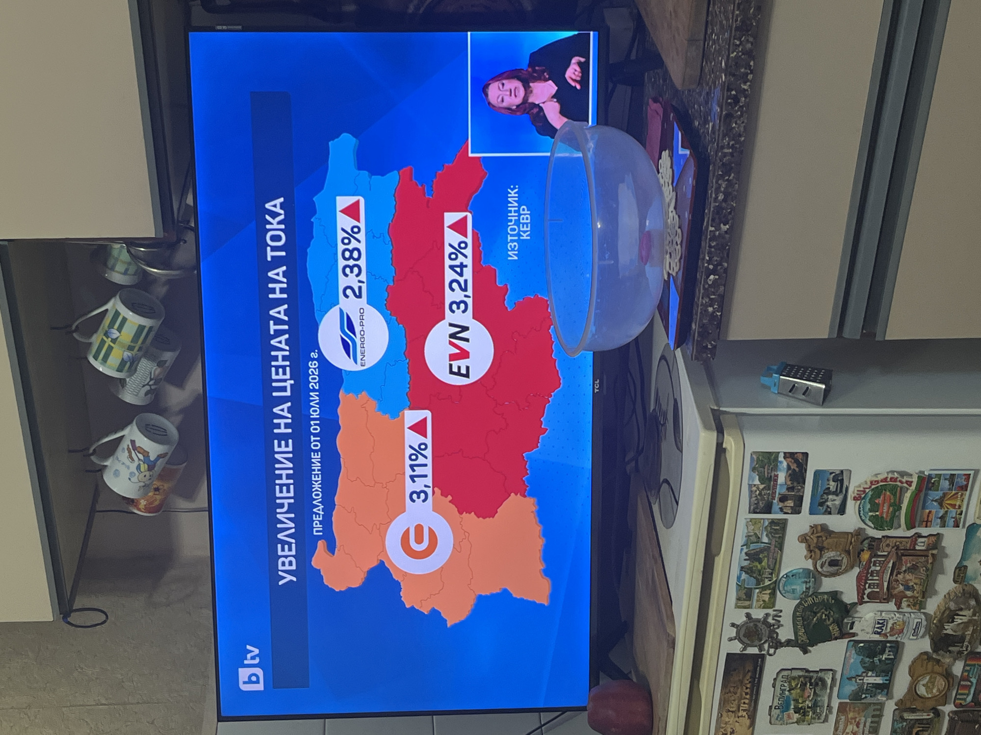 The image depicts an indoor scene, likely a kitchen, in the city of Burgas, Bulgaria. A television displaying a news broadcast is the central focus. The TV screen shows a graphical representation of Bulgaria's map, segmented into three regions indicating proposed electricity price increases by different providers, under the headline "УВЕЛИЧЕНИЕ НА ЦЕНАТА НА ТОКА" (Increase in electricity price). A subtitle reads "ПРЕДЛОЖЕНИЕ ОТ 01 ЮЛИ 2026 г." (Proposal from July 01, 2026).
The western orange region of the map displays a 3.11% increase with a generic electricity icon. The northeastern blue region shows a 2.38% increase attributed to "ENERGO-PRO." The southeastern red region indicates a 3.24% increase for "EVN." The source of this information is listed as "ИЗТОЧНИК: КЕВР" (Source: KEVR - Bulgarian Energy and Water Regulatory Commission). In the bottom right corner of the screen, a female sign language interpreter, dressed in a dark top, is actively performing sign language. The "bTV" channel logo is visible in the top left, and the television brand "TCL" is at the bottom center of the screen frame.
Above the television, light-colored kitchen cabinets are visible, with three assorted mugs hanging from hooks on the underside of a shelf. To the left of the screen, a red apple rests on a wooden cutting board. Below the TV, a dark, speckled countertop holds a transparent plastic bowl containing what appears to be seeds, placed on a blue and red coaster. To the right, a white refrigerator is covered with numerous souvenir magnets, many depicting various locations in Bulgaria, including one shaped like a miniature blue grater.