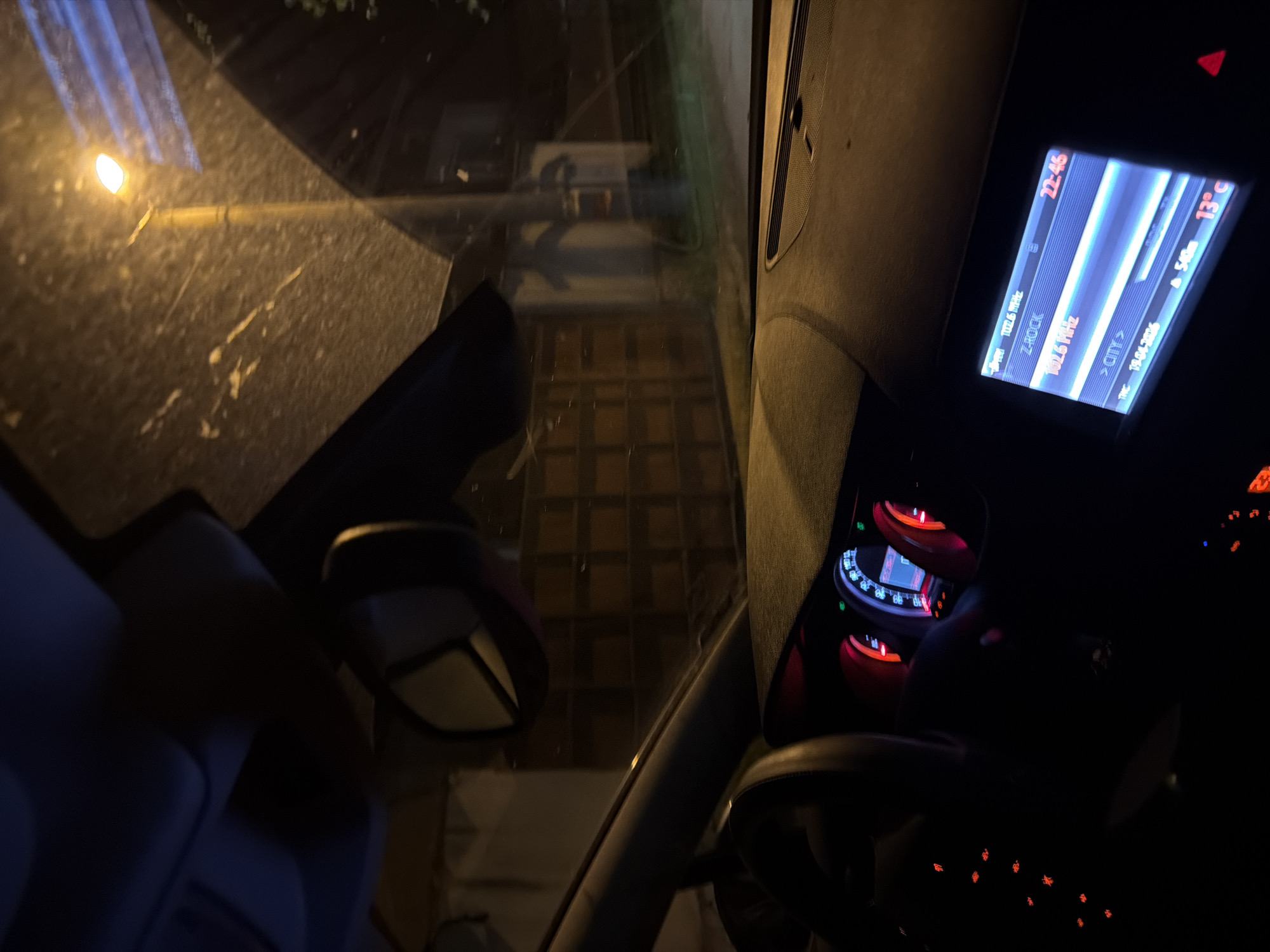 The image depicts the interior of a car at night. The dashboard and steering wheel are visible, with illuminated gauges and buttons. The radio display shows the time as 22:46, the frequency 102.6 MHz, and a temperature of 13°C. The car's side mirror is also visible. Through the windshield, a street with paving stones is seen, lit by a streetlight. The radio station named Z-ROCK suggests the location might be Sofia, Bulgaria, as the radio station's name and the city are displayed on the unit. The date TPC 1904-2016 is displayed on the radio.