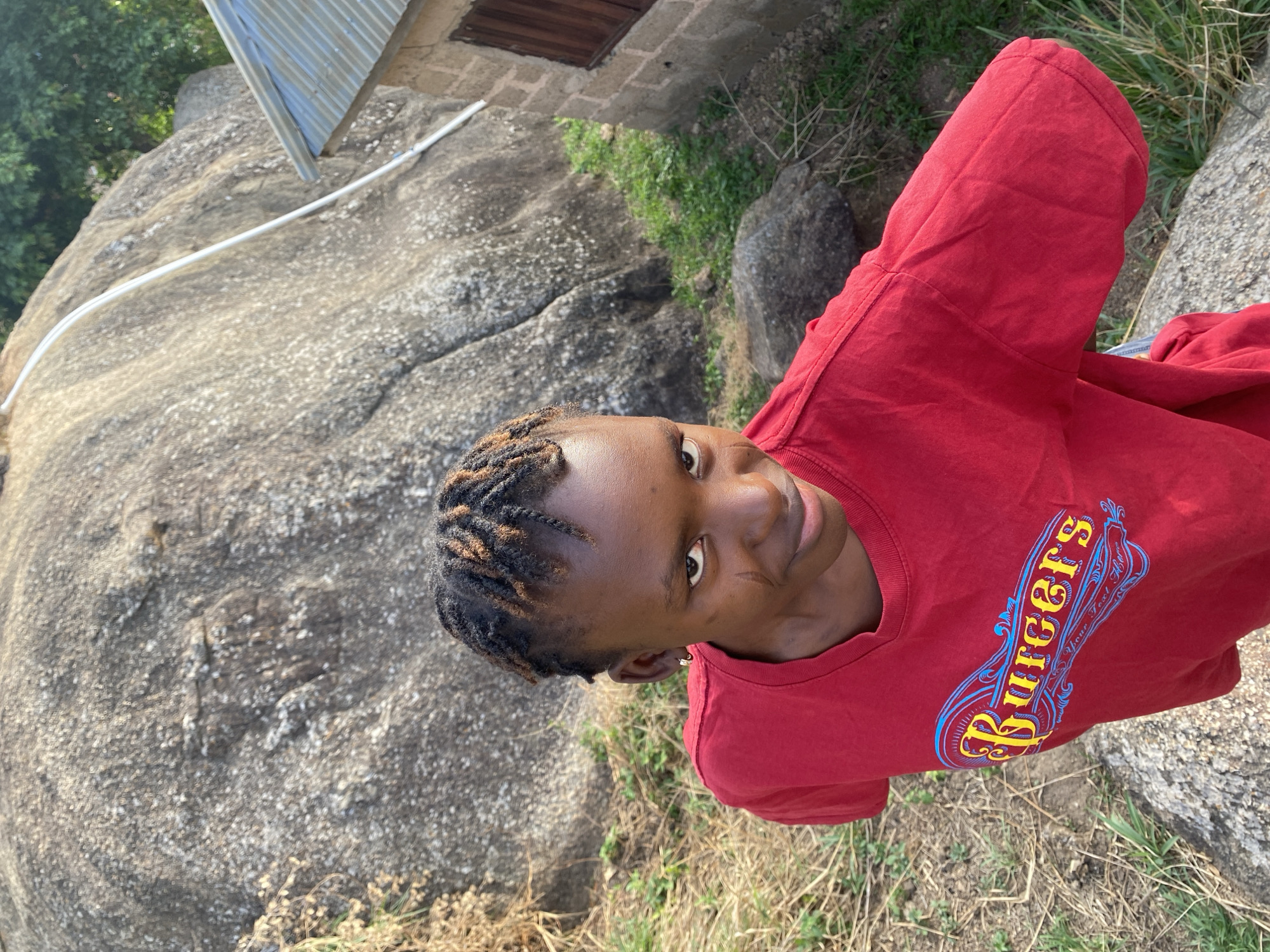 A young person is standing outdoors in Vom, Nigeria. They are wearing a red t-shirt with "Burgers" printed on it. The background includes a large rock formation with a metal roof structure attached to it, some grass, and building. The person appears to be looking at the camera.