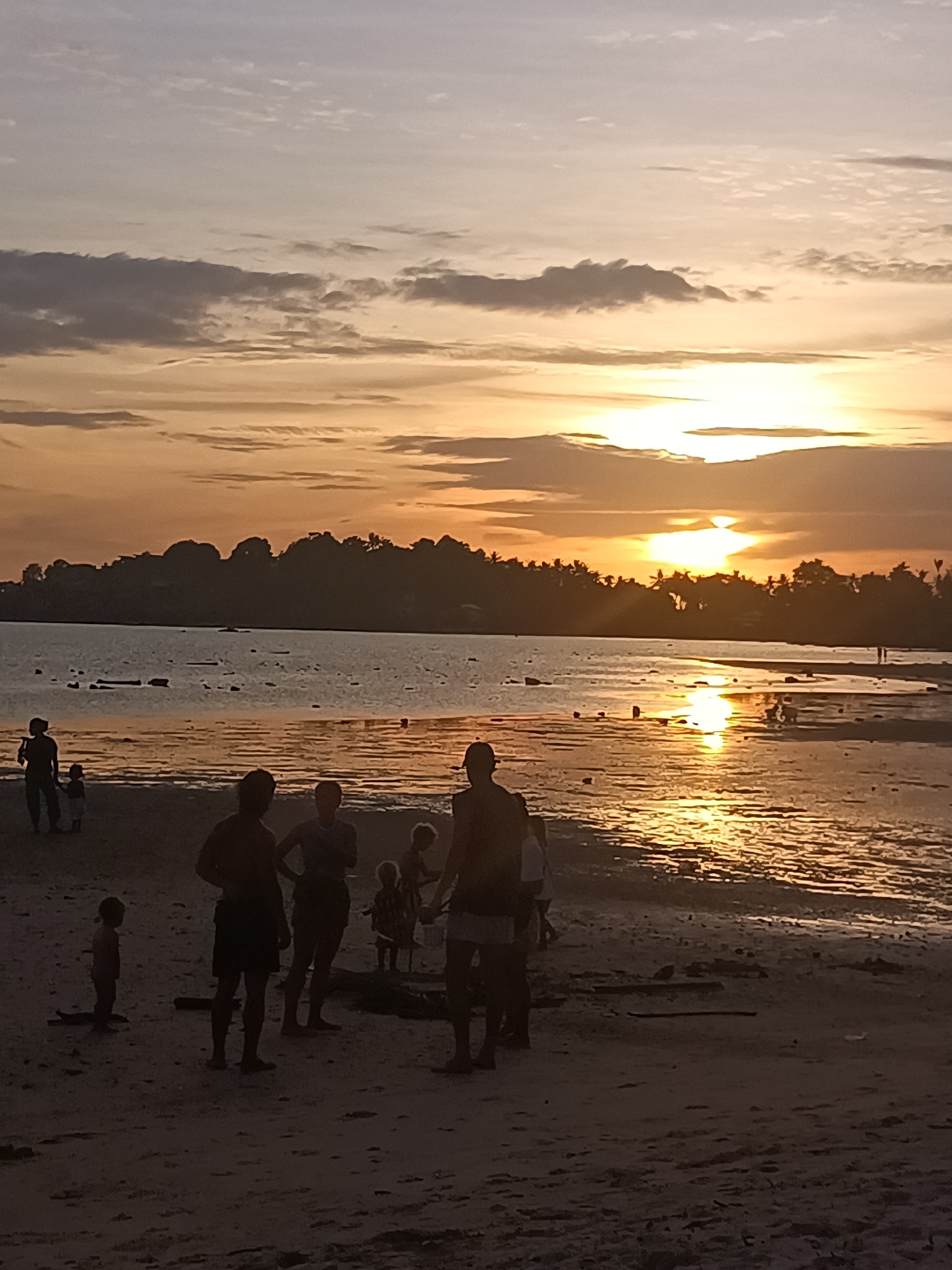 A group of people are gathered on a beach in front of a body of water during sunset. The sky is filled with hues of orange, yellow, and gray clouds, with the sun low on the horizon reflecting on the water. Silhouettes of people are visible on the sand, some standing, some interacting, and one child standing alone. In the background, a tree-lined silhouette of land is visible across the water. The sand appears wet in some areas, suggesting the tide is out or has recently receded. The scene depicts a tranquil evening on the coast of Koh Phangan, Thailand.