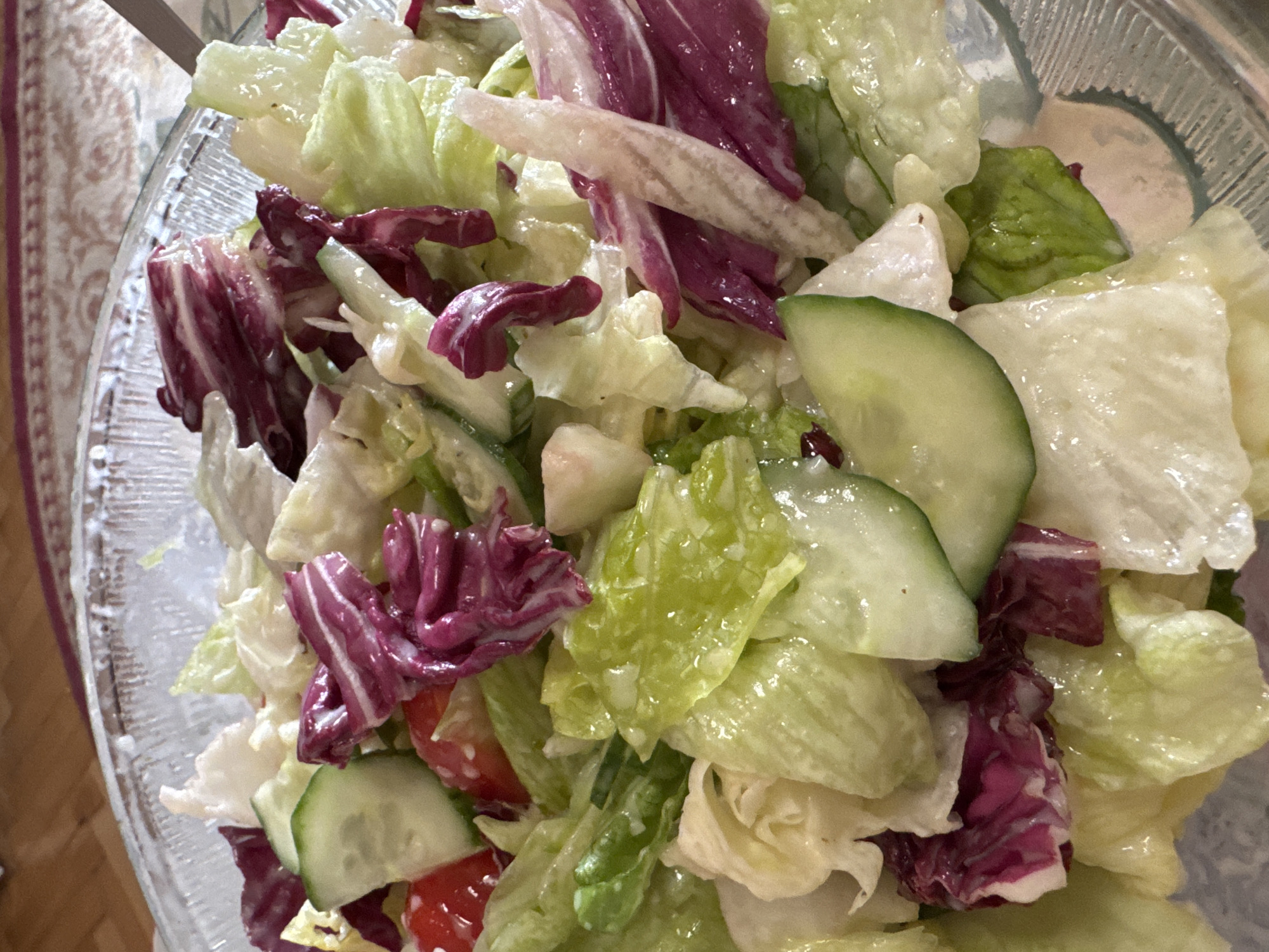 A close-up shot of a salad in a glass bowl. The salad contains various types of lettuce, including iceberg and radicchio, cucumber slices, and red tomato pieces, all coated in a creamy dressing. The background is slightly blurred, showing a table with a patterned cloth. The image does not show location information (Sofia, Bulgaria).