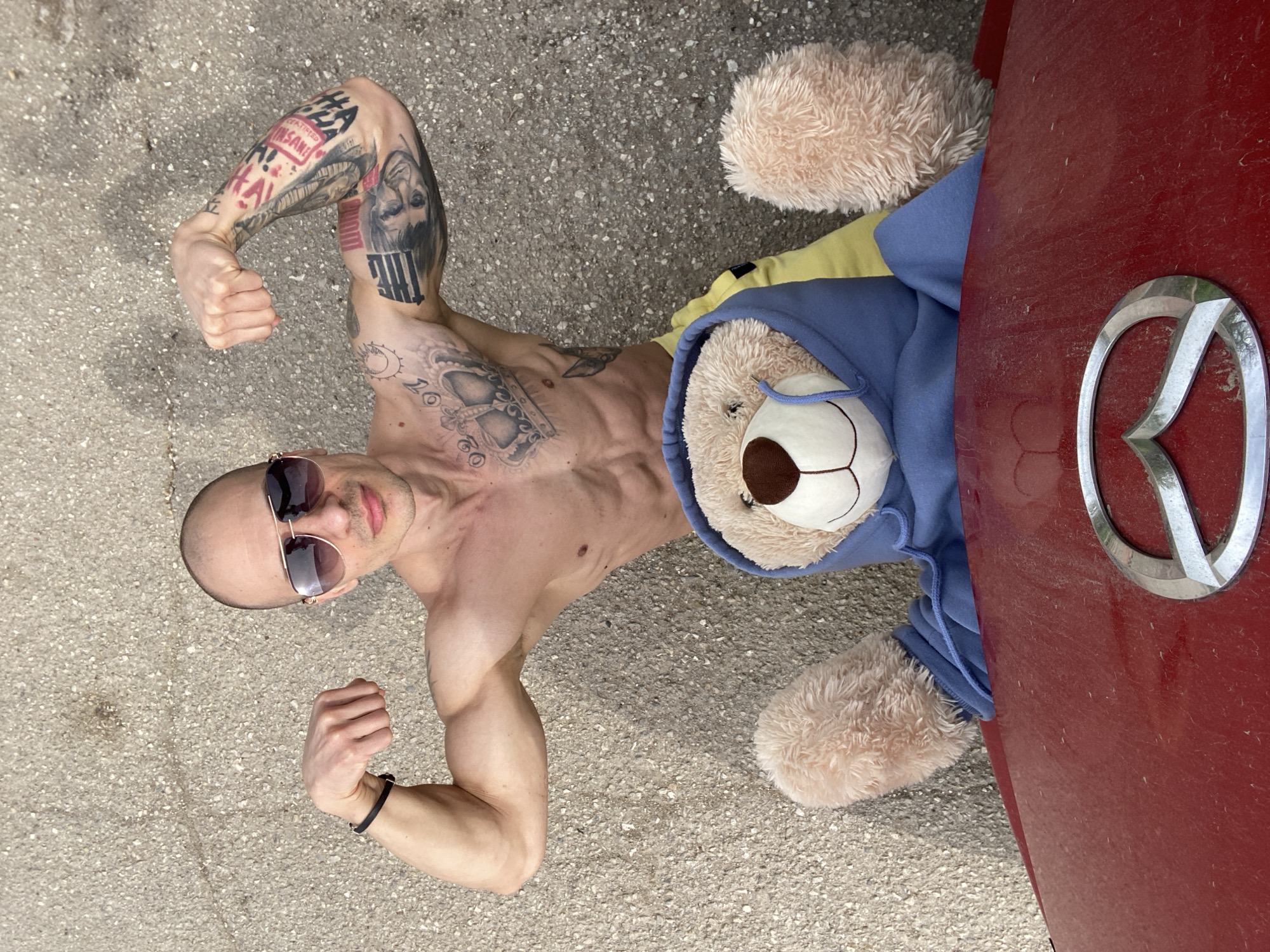 The image presents an overhead view of a shirtless man lying on asphalt. The man is muscular and tattooed, wearing sunglasses and a bracelet. He is flexing his arms. A teddy bear wearing a blue hooded sweatshirt and a yellow shirt lies partially under the red hood of a Mazda car. The location is implied to be in the city of Pleven, Bulgaria.