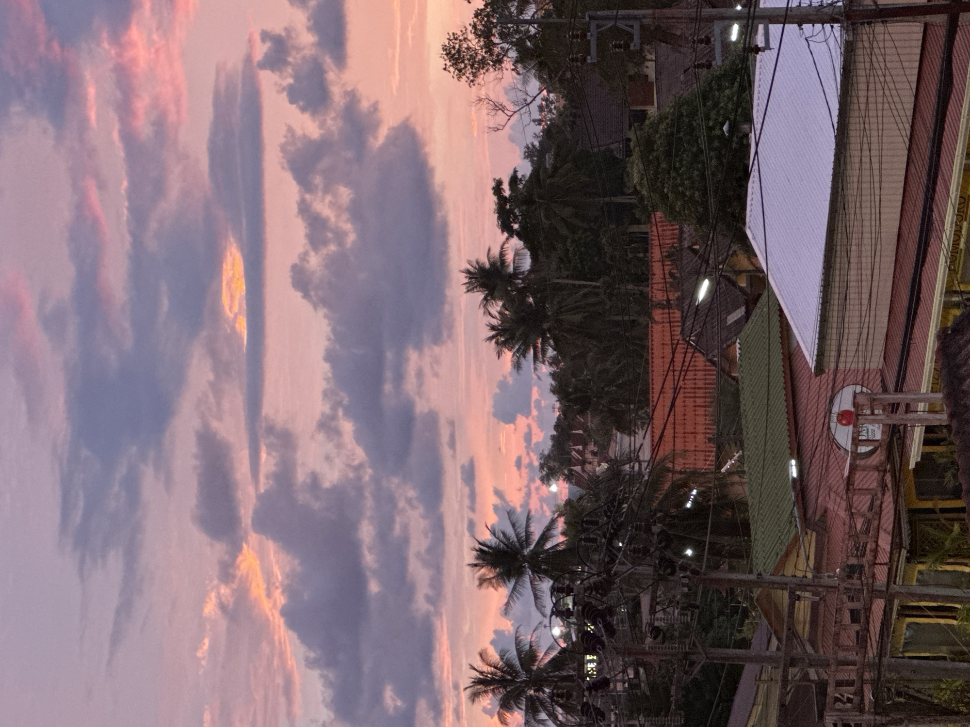 This media file captures a picturesque sunset over a tropical town, likely Karon, Thailand. The sky is a canvas of soft pinks and purples, streaked with dramatic grey clouds that catch the fading sunlight, creating pockets of vibrant orange. This vibrant sky suggests a beautiful evening, with the soft colors evoking a sense of peace and tranquility.
Below the sky, the scene transitions to a bustling street lined with buildings and lush vegetation. Palm trees with their distinctive fronds dot the landscape, their silhouettes sharp against the colorful sky. Various buildings, some with red-tiled roofs and others with corrugated metal sheeting in green and white, are visible. The architecture suggests a tropical climate and a developed area, possibly a commercial or residential district.
Numerous utility poles and wires crisscross the scene, indicating an active community. Streetlights are also visible, some illuminated, hinting that dusk is settling in. A notable object in the mid-ground is a round sign with a red circle and what appears to be text, possibly a business logo, on a pink building. The overall impression is one of a vibrant, tropical town experiencing the serene beauty of a sunset. The time of day is clearly sunset, given the colors of the sky and the presence of artificial lights. The weather appears clear for the sunset to be so vivid.