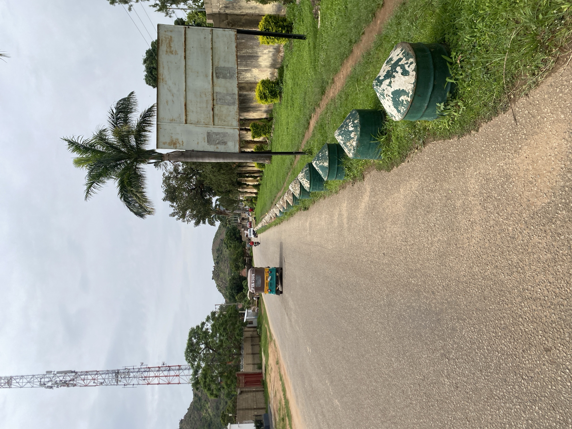 In Jos, Nigeria, on a cloudy day, a paved road curves slightly to the left. A three-wheeled taxi, with the license plate "FTA-2984", travels down the road, heading away from the viewer. The taxi is primarily yellow, with a blue and green section at the rear. A person is visible in the driver's seat. On the right side of the road, a grassy verge is lined with several conical, green-and-white painted bollards. Behind the bollards, a weathered concrete wall runs along the edge of a property. A large, blank billboard stands near the wall, partially obscured by a palm tree. Further down the road, other vehicles and people are visible, along with trees and rolling hills in the background. A tall telecommunications tower, red and white, stands on the left side of the road, near a fence. The scene suggests a typical streetscape in Jos, characterized by its moderate climate, local transportation, and the presence of communication infrastructure.