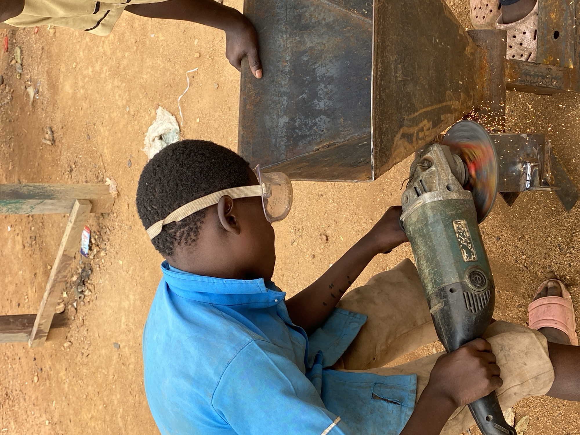 An overhead shot captures a young person, likely a child or teenager, in a blue shirt and khaki pants, performing metalwork outdoors in Kuru Dungus, Nigeria. The individual wears clear safety goggles and operates a green and black angle grinder with both hands, directing it towards a dark metal workpiece. Bright orange and red sparks fly vigorously from the grinding disk, indicating active cutting or shaping of the metal.
Part of another person's arm and hand are visible at the top of the frame, holding the metal workpiece steady for the grinder operator. To the lower right, a foot wearing a pink slide sandal is partially visible. The setting appears to be an unpaved outdoor workshop or construction area, with the ground composed of light brown dirt or sand, scattered with small stones, wood debris, and what looks like a piece of white fabric. On the side of the angle grinder, some faded text or a logo is visible, which appears to be "SUPER" or a similar word. The scene depicts a manual labor activity taking place under bright daylight conditions.