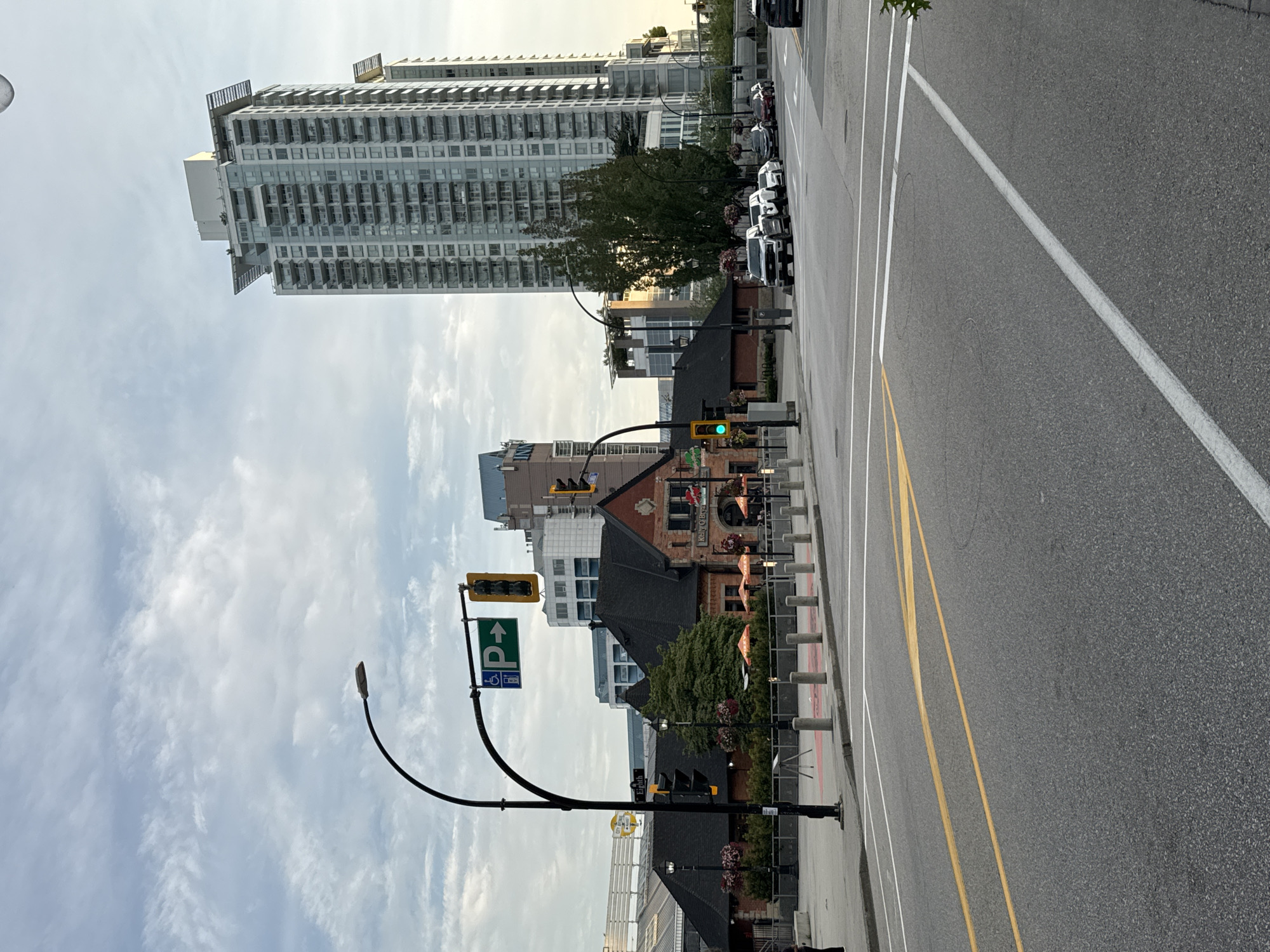 This is a street-level photograph taken in the city of New Westminster, Canada, looking southeast on a cloudy day. The foreground features an asphalt road with yellow and white lane markings. A crosswalk is visible on the right side of the image. In the middle ground, a red brick building with a dark roof and awnings is situated, identified by signage as "Kelly O'Bryan's". Adjacent to this building is a pedestrian walkway with bollards and planters. A tall, modern glass-and-steel skyscraper dominates the background, with several other buildings of varying heights also present. Traffic lights and streetlights are visible throughout the scene. Several vehicles are parked or stopped along the street. The sky is overcast with scattered clouds.