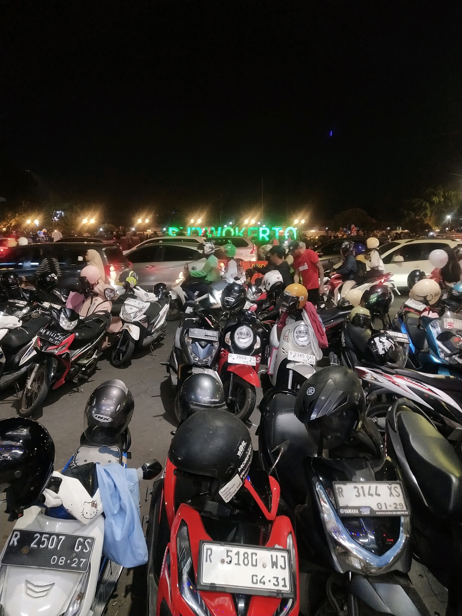 The image captures a nighttime scene in Purwokerto, Indonesia, characterized by a densely packed parking lot filled with numerous motorcycles. In the background, bright green neon lights spell out "PURWOKERTO," illuminating the dark sky. Several cars are also visible, parked amongst the motorcycles. A mix of individuals, some wearing helmets and others without, are present, with some appearing to be interacting with the parked vehicles. The environment is outdoors, with artificial lighting from street lamps and the neon sign creating a vibrant, yet somewhat chaotic, atmosphere. The foreground is dominated by the close-up view of several motorcycles and their helmets.