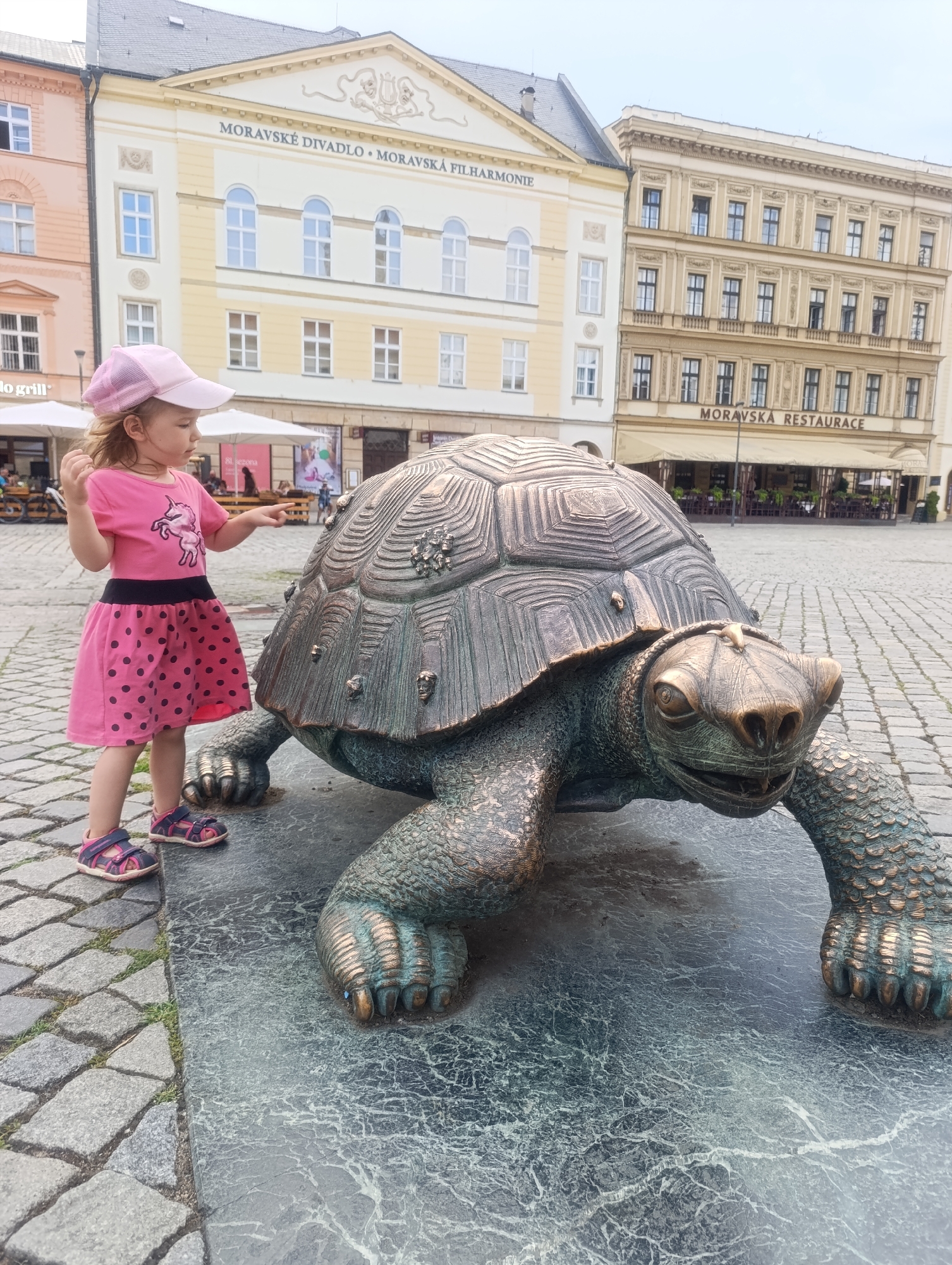 In Olomouc, Czechia, a young girl wearing a pink t-shirt with a unicorn graphic and a polka-dotted skirt, and a pink cap, stands on a cobblestone plaza next to a large bronze turtle sculpture. The sculpture appears to be a public art installation, possibly a landmark. In the background, a building with the inscription "MORAVSKÉ DIVADLO • MORAVSKÁ FILHARMÓNIE" is visible, suggesting the location is near a theater and philharmonic hall. Another building to the right has a sign that reads "MORAVSKÁ RESTAURACE". The sky is overcast, and the scene suggests an outdoor urban environment.