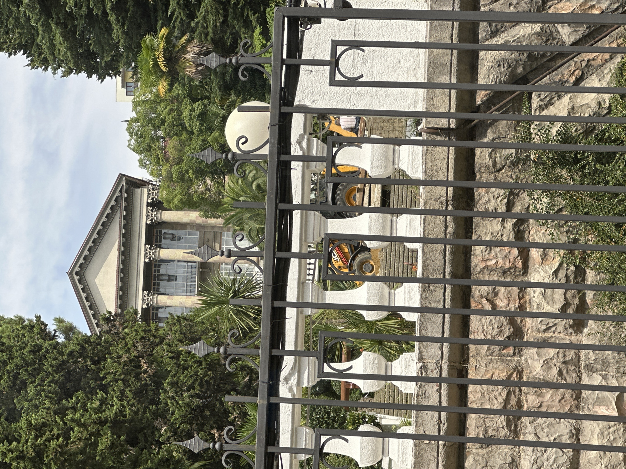 In Sochi, Russia, the image captures a view through a dark metal fence, partially obscuring a white balustrade with decorative elements. Behind the balustrade, a yellow construction vehicle, likely a backhoe loader with the branding "JCB" visible, is partially obscured by greenery. Further back, a large, classical building with columns and a pediment is visible amidst dense trees and palm trees. The sky above is overcast.
