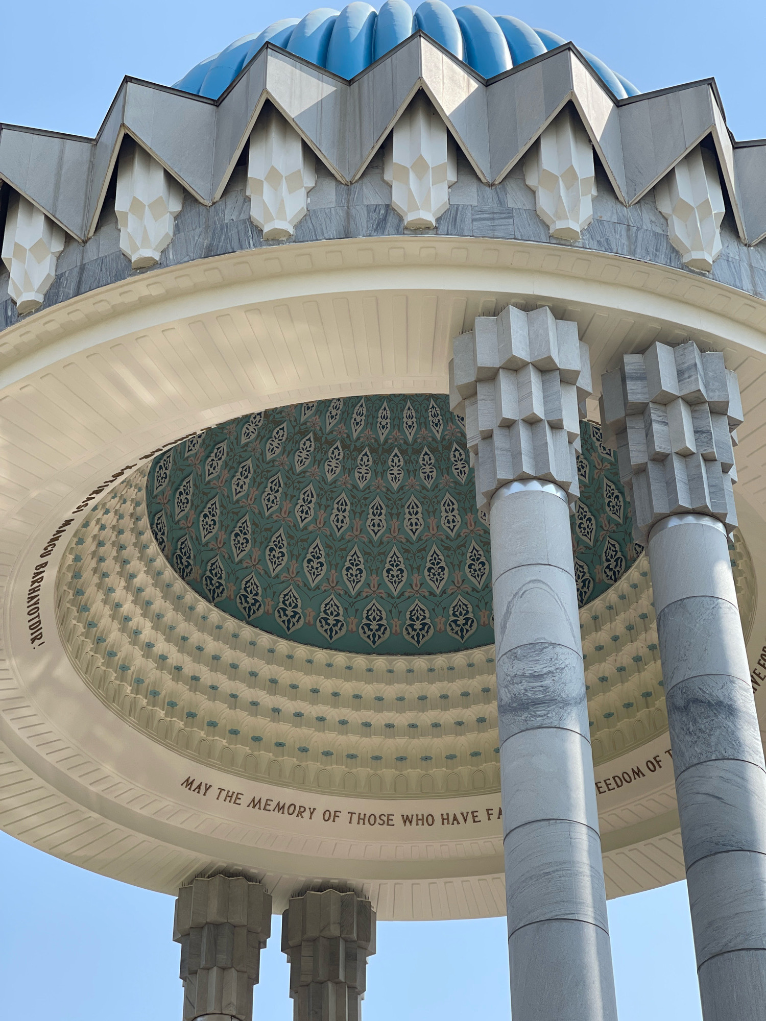 This is a low-angle, outdoor, eye-level shot of the exterior of the Independence Monument in Tashkent, Uzbekistan, under a clear blue sky. The monument features a prominent blue dome with a fluted design, topped by a golden finial. A decorative frieze with triangular grey elements and white geometric carvings runs around the base of the dome, above a band of grey marble. Below this, a white, curved structure with ornate white and light blue decorative elements encircles the interior.
Two large, cylindrical marble columns with stacked geometric stone blocks at the top support the white structure. The interior of the dome is visible, adorned with a repeating pattern of intricate teal and dark blue floral motifs. Inscriptions are present on the white encircling structure. On the left, in darker lettering, it reads "BARHAYOTDIR!". On the right, also in darker lettering, it reads "FREEDOM OF T". Partially visible text below this suggests "MAY THE MEMORY OF THOSE WHO HAVE FA".
There are no people visible in the image. The scene is static and focuses solely on the architectural details of the monument. The lighting suggests it is daytime, with shadows cast by the architectural elements.
