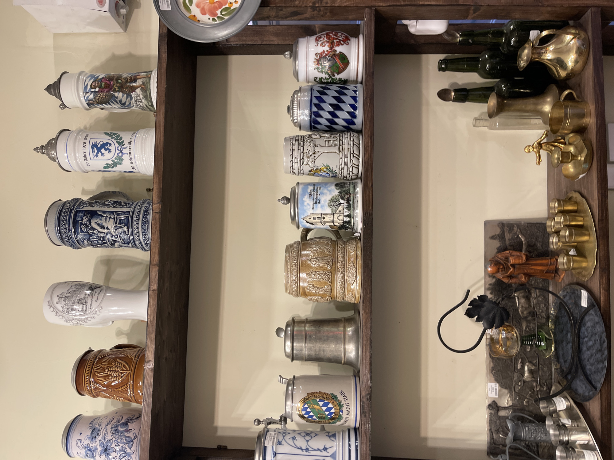 This image shows a close-up of several shelves in what appears to be a store or a private collection. The shelves are made of dark wood and are filled with various decorative items.
On the top shelf, there are several ceramic beer steins. From left to right, there is a white stein with blue floral patterns, a brown stein with wheat imprints, a white stein with a landscape depiction, and two more ceramic steins, one with a blue crest and text reading "20 Jahre 1976-1996 FC Scheuerrer", and another with a man in historical attire. Further to the right is a metallic object with floral decorations.
The middle shelf contains a variety of items. On the left, there is a tall blue and white ceramic mug. Next to it is a lighter colored ceramic mug with a coat of arms and text. Then there is a metallic lidded container, followed by a brown ceramic stein with intricate relief carvings of figures and foliage. To the right of this are two more ceramic steins, one with a depiction of a church and trees, and another with a blue and white diamond pattern, and a final stein with a coat of arms.
The bottom shelf holds a collection of various objects. On the far left, there are stacks of small metallic cups. In the center, there is a decorative dark tray with a small glass resting in a metal holder shaped like a vine, and to the right of this is a figurine of a monk. Further to the right, there is a tray with small brass cups, a brass candlestick with a small figurine, a small brass mug, and a larger brass pitcher or ewer. Behind these items, there are several dark green glass bottles and some clear glass bottles.
The background is a plain, light-colored wall. The lighting suggests an indoor environment, possibly with artificial light. The overall impression is of a display of collectible items, likely related to German culture given the presence of beer steins. The location is stated to be Svetlogorsk, Russia.