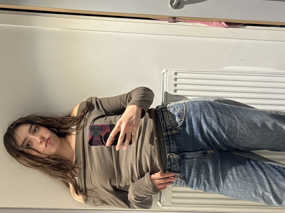 A young woman with long brown hair poses, holding a cell phone with a colorful case, in front of a mirror. She wears a brown, long-sleeved shirt with shoulder cutouts and blue jeans. A white radiator is visible behind her jeans. Above the woman, the top of a door with a silver handle can be seen. A pink item peeks out from the space between the door and the frame. The background is a plain white wall.