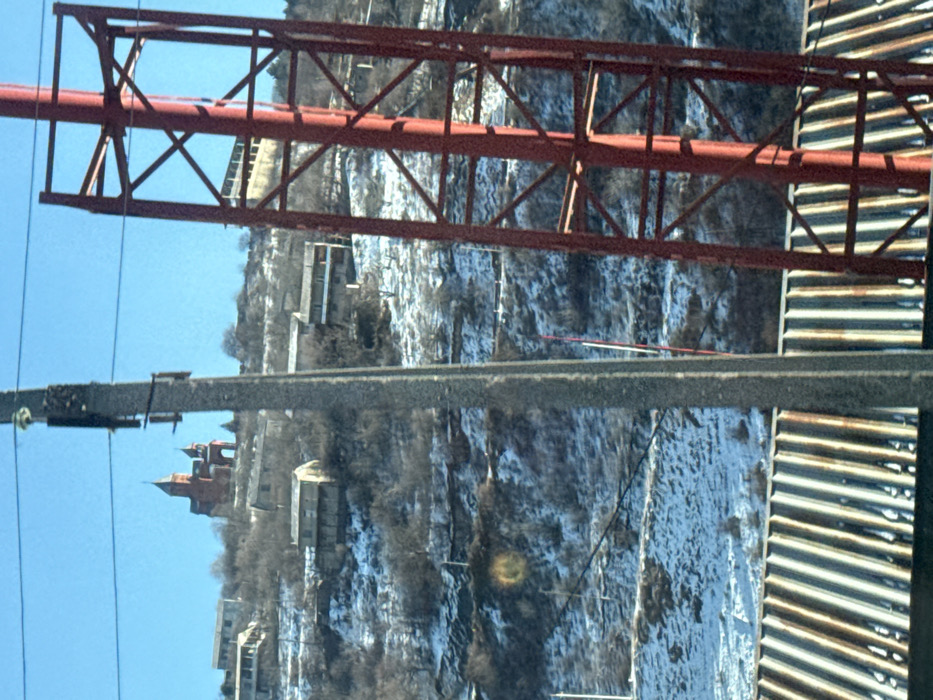 The image displays a panoramic view of a snow-dusted hillside in the city of Koghb, Armenia, under a clear blue sky. In the foreground, the top of a corrugated metal roof is visible at the bottom of the frame. Obscuring parts of the scene are a grey concrete utility pole positioned slightly left of center, extending vertically upwards, and a red metal lattice tower on the right, partially visible. Multiple thin power lines traverse the upper portion of the frame.
The midground and background depict a steep, terraced hillside partially covered with patches of snow and bare deciduous trees or shrubs, suggesting a winter or early spring season. Numerous residential structures, primarily multi-story buildings with light-colored facades and dark roofs, are scattered across the slope.
Dominating the highest point of the hill in the background is a distinctive red architectural structure, identified as the Surb Amenaprkich (Holy All-Savior) Church, which features multiple conical domes and a prominent central dome. No individuals or active interactions are observed in the scene.