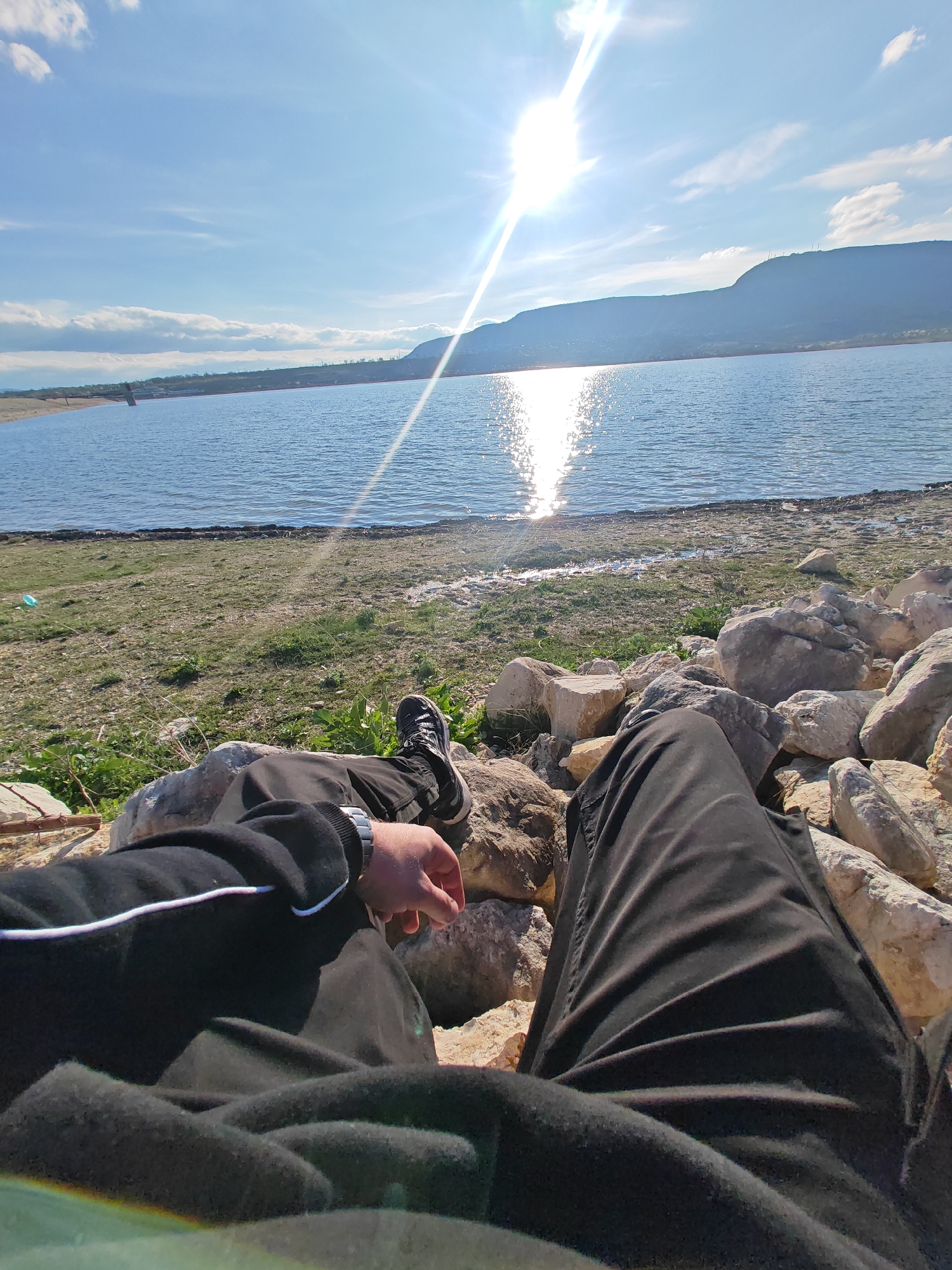 The image captures a first-person perspective of someone relaxing by a large body of water on a bright, sunny day. The lower half of the frame is dominated by the person's legs, clad in dark, possibly black, casual pants, and their left arm wearing a similar dark long-sleeved top with a thin white stripe near the cuff. A dark-colored watch is visible on the left wrist. The person appears to be sitting among an array of light-colored, greyish rocks and boulders that form a rocky shoreline or embankment.
Beyond the immediate foreground, a narrow strip of sparse, dry green grass and patches of bare earth stretches towards the water's edge. The main feature of the midground is a vast expanse of blue water, likely a lake or large reservoir, with gentle ripples visible across its surface. A brilliant sun glare creates a shimmering path of light stretching from the horizon directly towards the viewer on the water's surface, indicating the sun's position.
In the background, a distant, hazy mountain range or hills line the horizon, appearing slightly desaturated due to the distance and atmospheric perspective. The sky above is a clear, bright blue with scattered white, fluffy clouds. The sun itself is prominently visible in the upper right quadrant of the image, emitting strong, visible rays of light and a lens flare effect. The overall scene suggests a tranquil outdoor setting, with the person enjoying the natural landscape and bright weather. There is no visible text in the image.