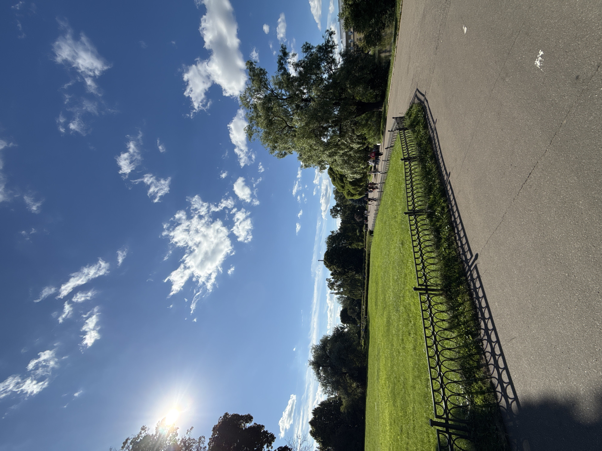 This is an outdoor, eye-level, wide-angle photograph of a park on a sunny day. The sky is a vibrant blue with scattered white clouds, and the sun is shining brightly, creating lens flare on the left side of the image. In the foreground, a paved pathway curves to the right. To the left of the path is a lush green lawn bordered by an ornate black metal fence. Beyond the lawn, various trees are visible, including a large, leafy tree on the right side of the frame. In the middle ground, a group of people are walking along the pathway, some appearing to be cycling. In the background, more trees and foliage can be seen, and a body of water is partially visible on the far right. A building with a light-colored facade is also visible in the far background on the right. The scene depicts a public park, likely in Moscow, Russia, given the architectural style of the building and the general aesthetic of the park.